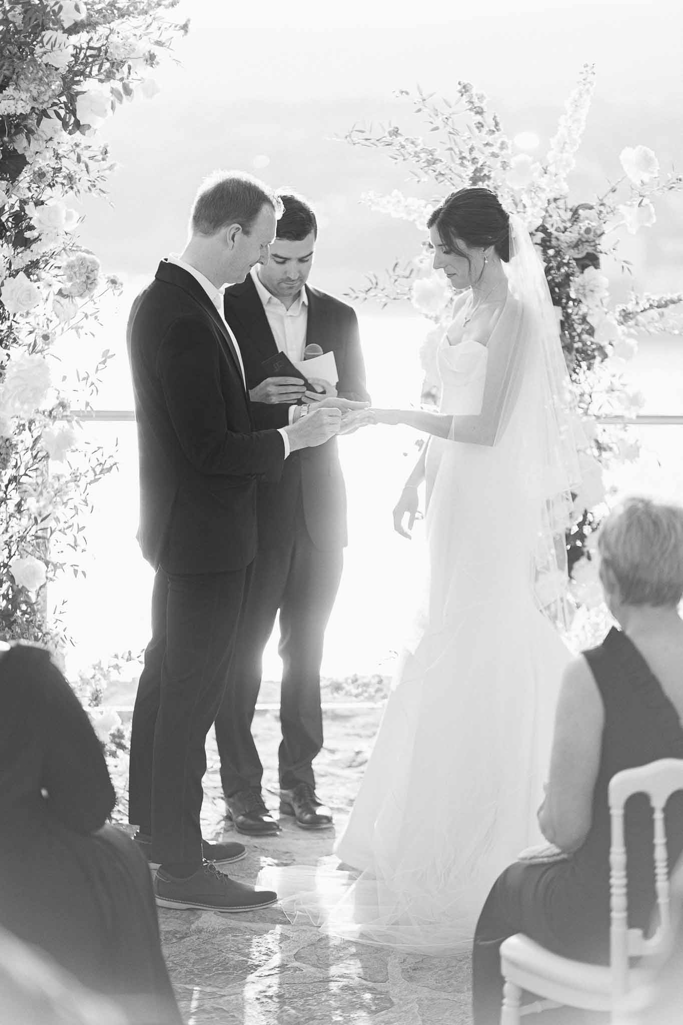 Wedding ceremony ring exchange with bride, groom and officiant at indoor venue with floral arch