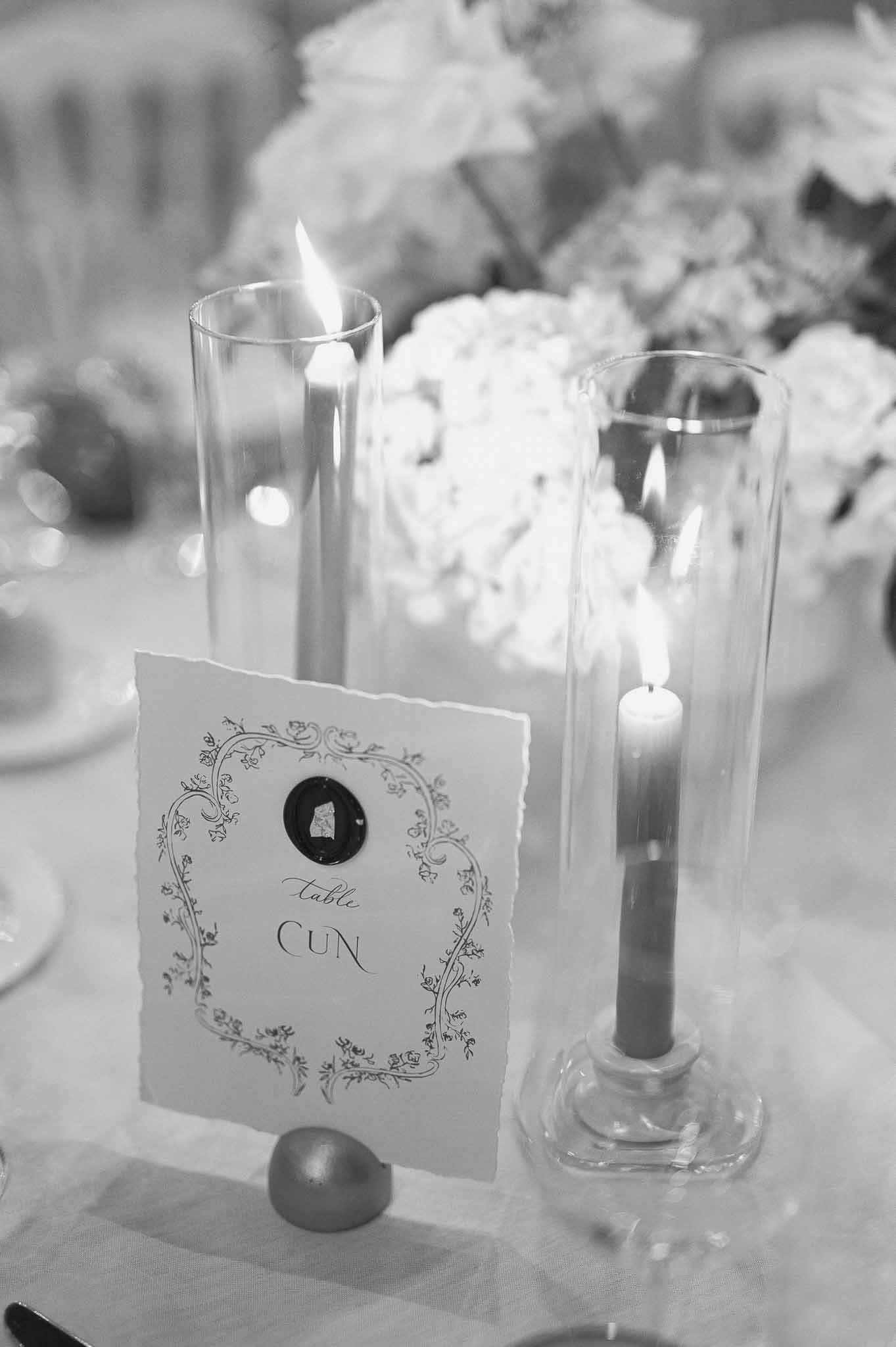Black and white close-up of formal table setting with place card, candles, and floral arrangements at reception