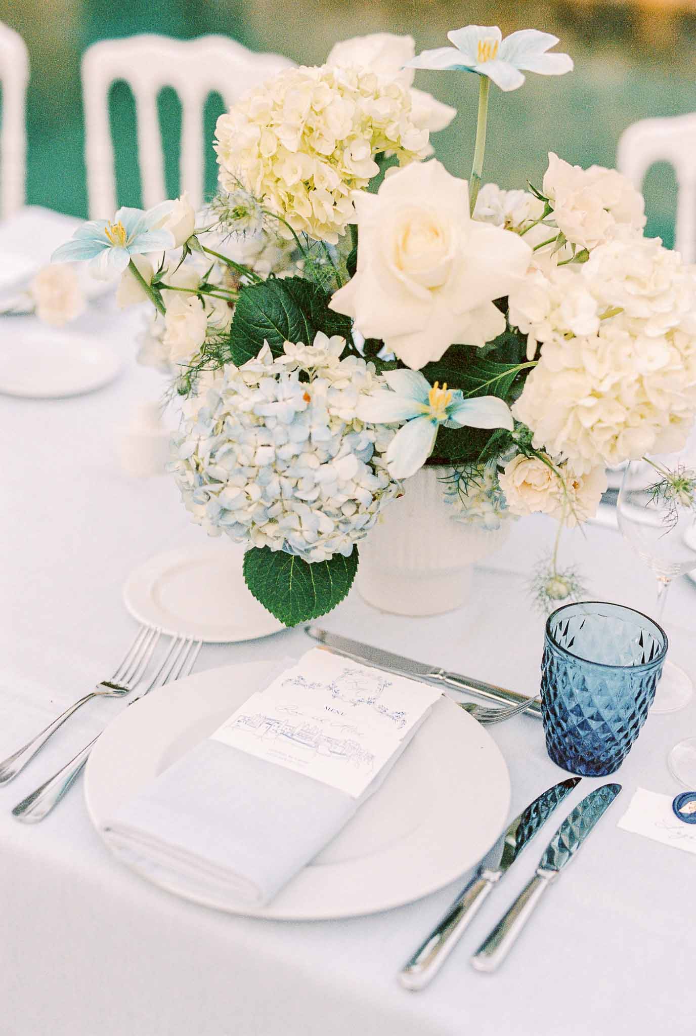 Wedding reception table setting with white floral centerpiece and blue glassware at outdoor garden venue