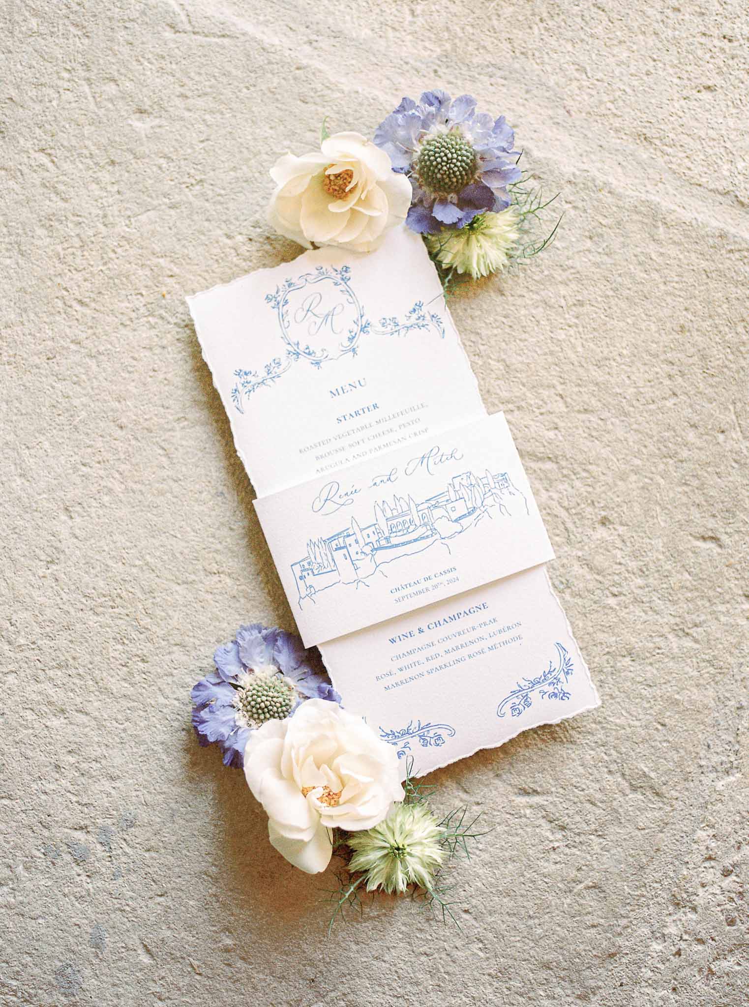 Wedding menu card with blue text and floral arrangements on stone surface at French château