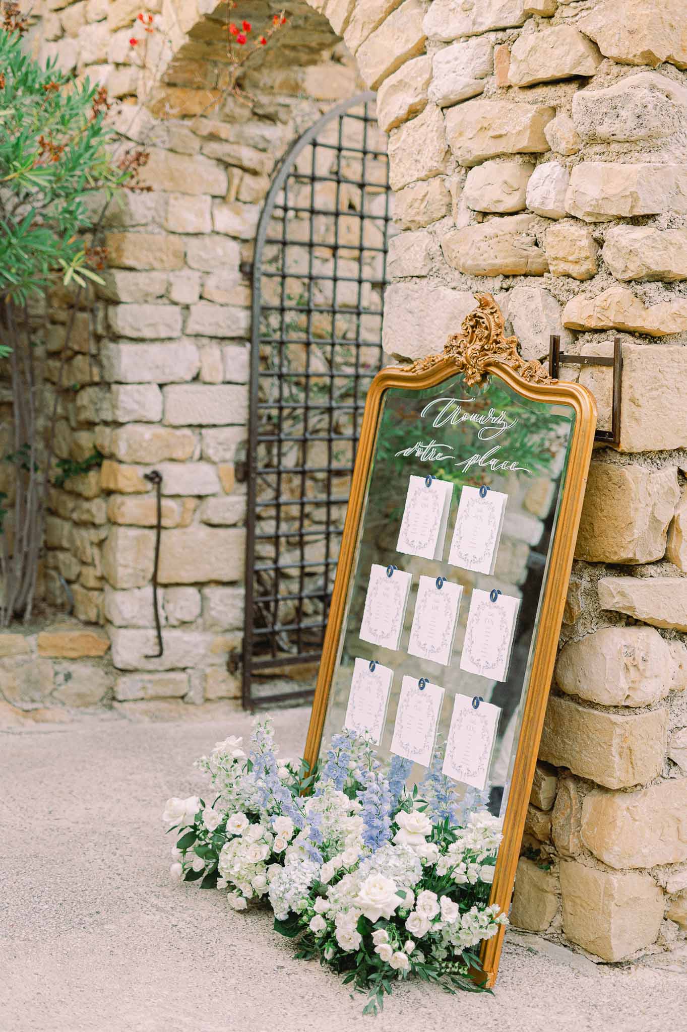 Gold-framed seating chart mirror with white roses and blue delphiniums at stone courtyard wedding venue