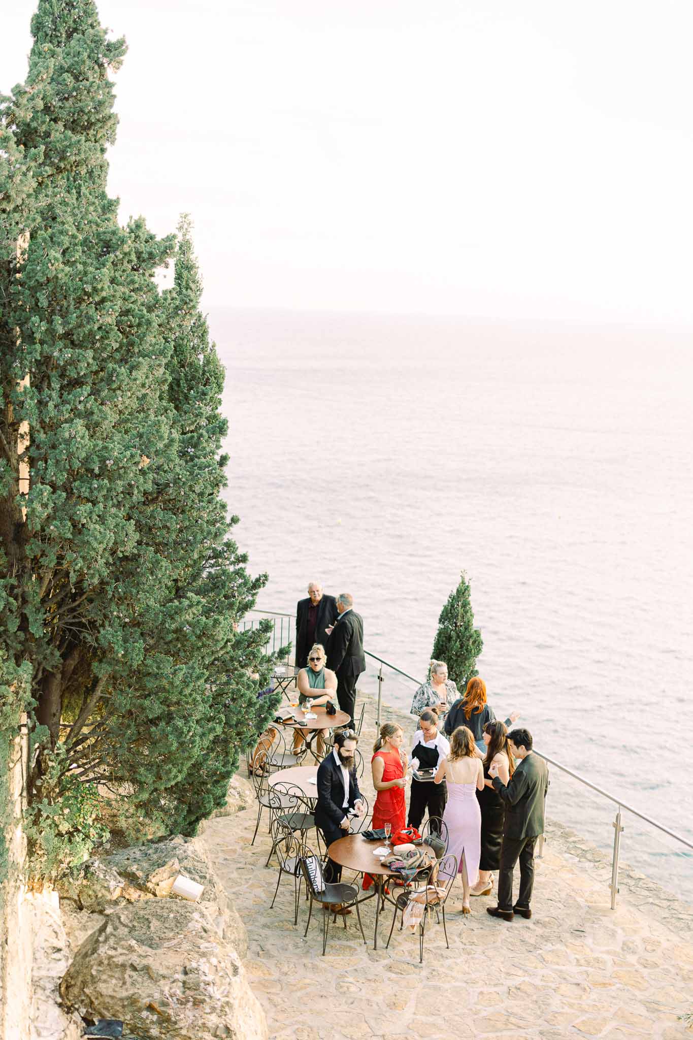 Cocktail hour on cliffside terrace with cypress trees overlooking water at Mediterranean venue