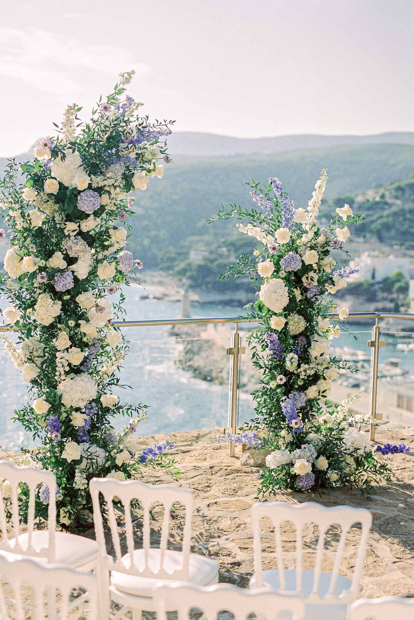 Outdoor wedding ceremony setup on clifftop overlooking water and mountains with floral arch and white chairs