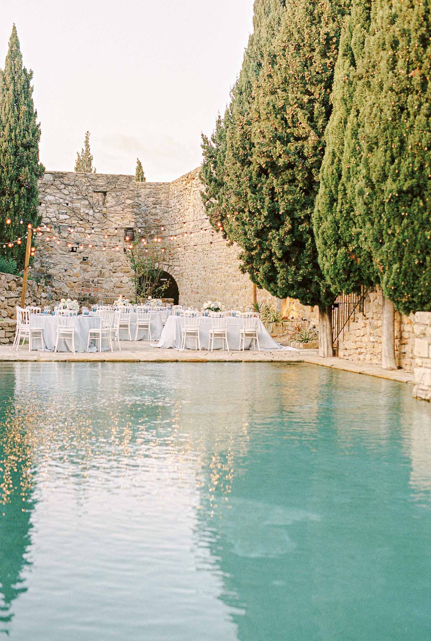 Outdoor wedding reception setup beside pool at historic stone fortress venue with cypress trees