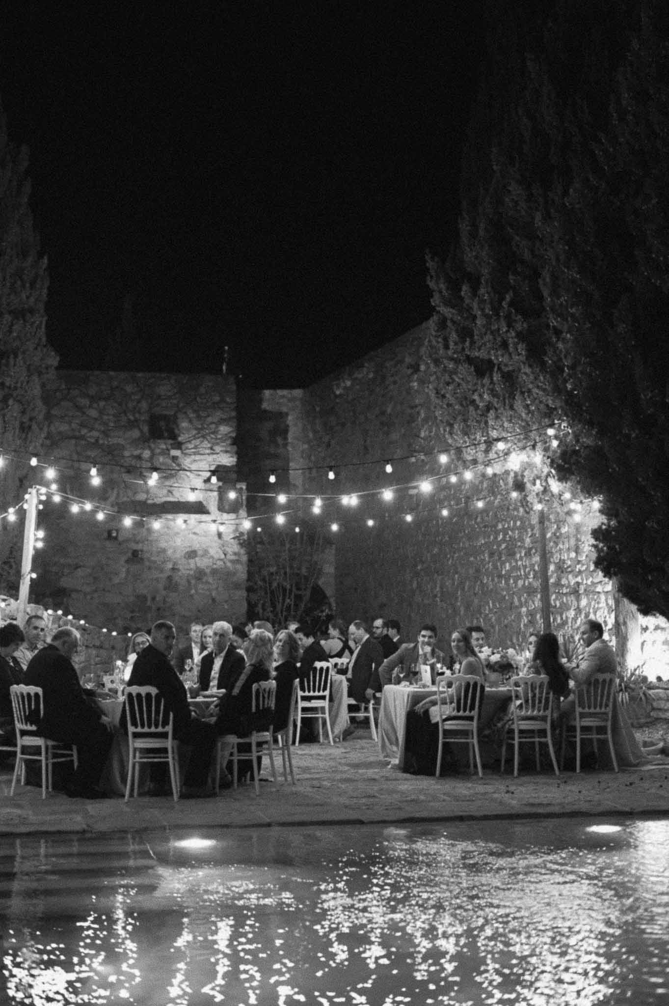 Evening reception with guests dining at tables in historic stone courtyard beside water