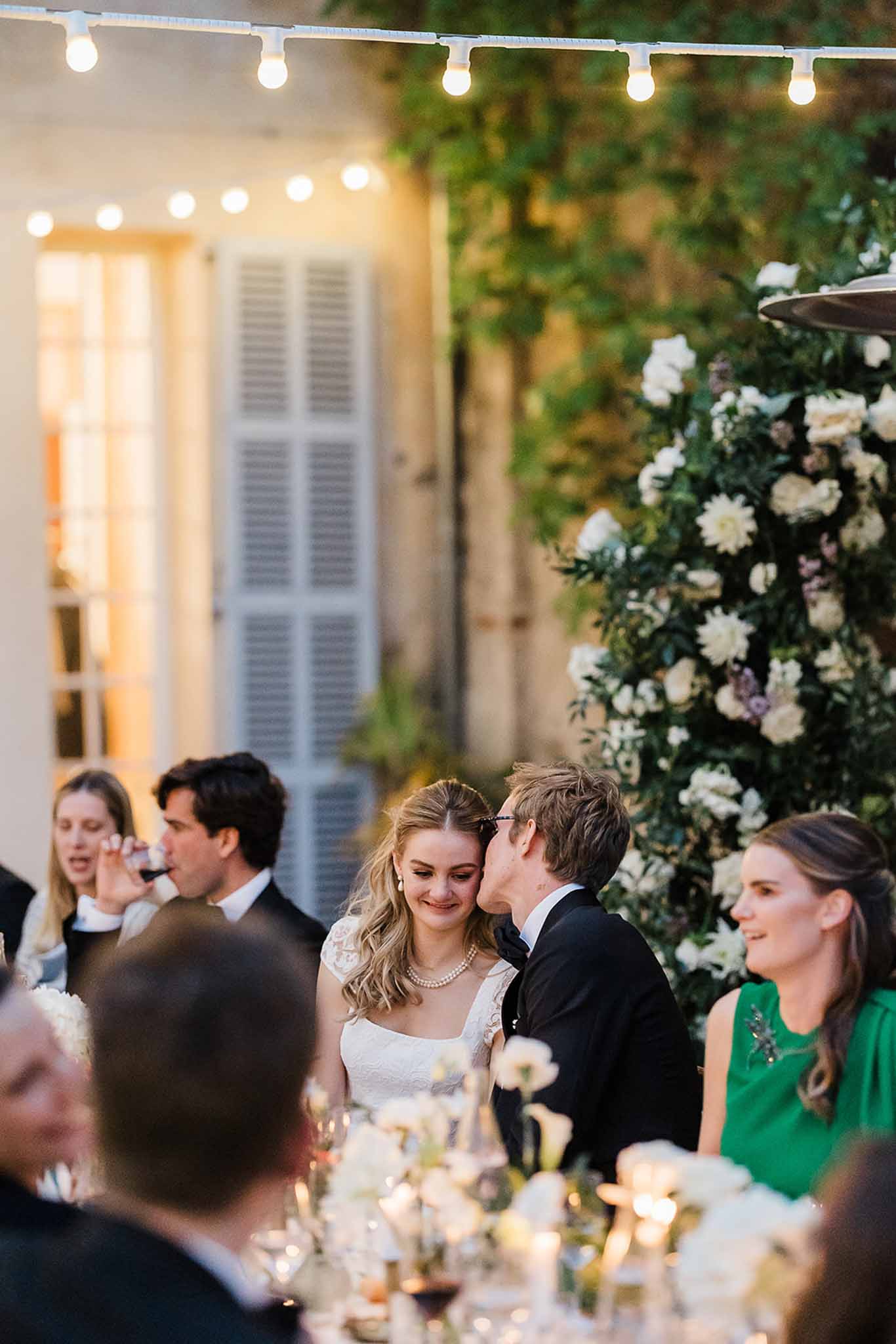 Bride and groom sharing tender moment during outdoor courtyard reception with floral arrangements and string lights