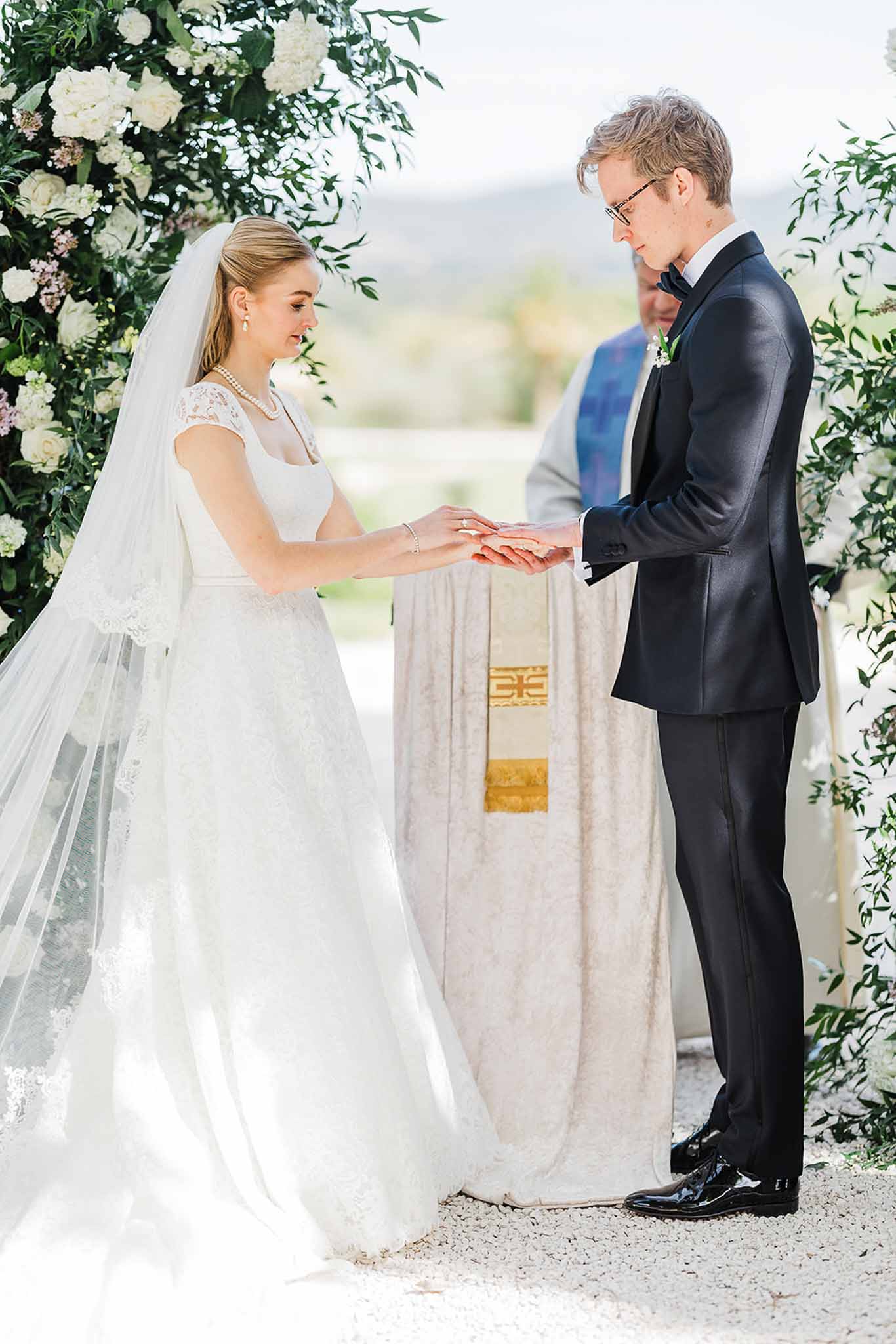 Bride and groom exchanging rings during outdoor wedding ceremony beneath floral arch