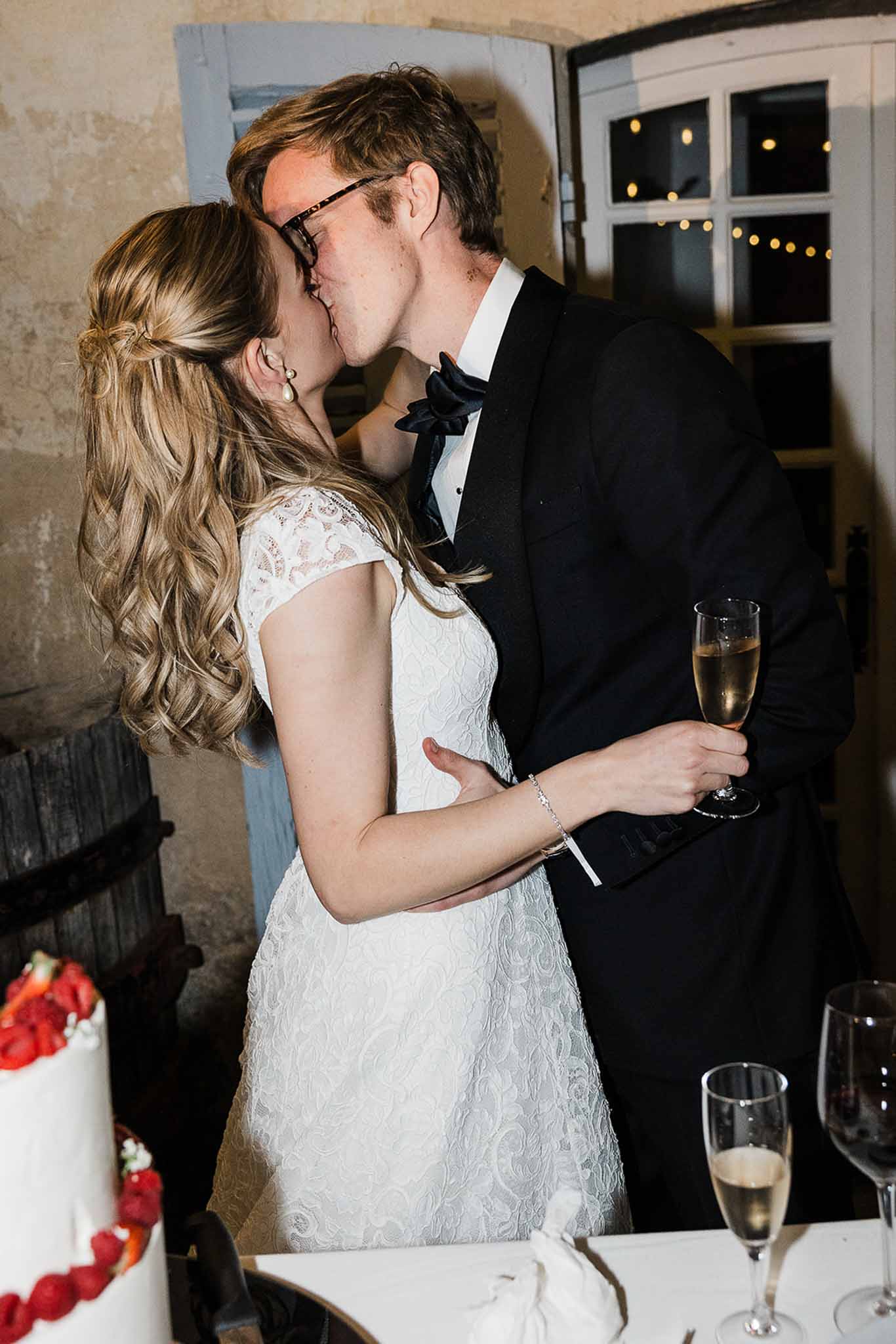 Bride and groom kissing with champagne glasses during indoor wedding reception with cake