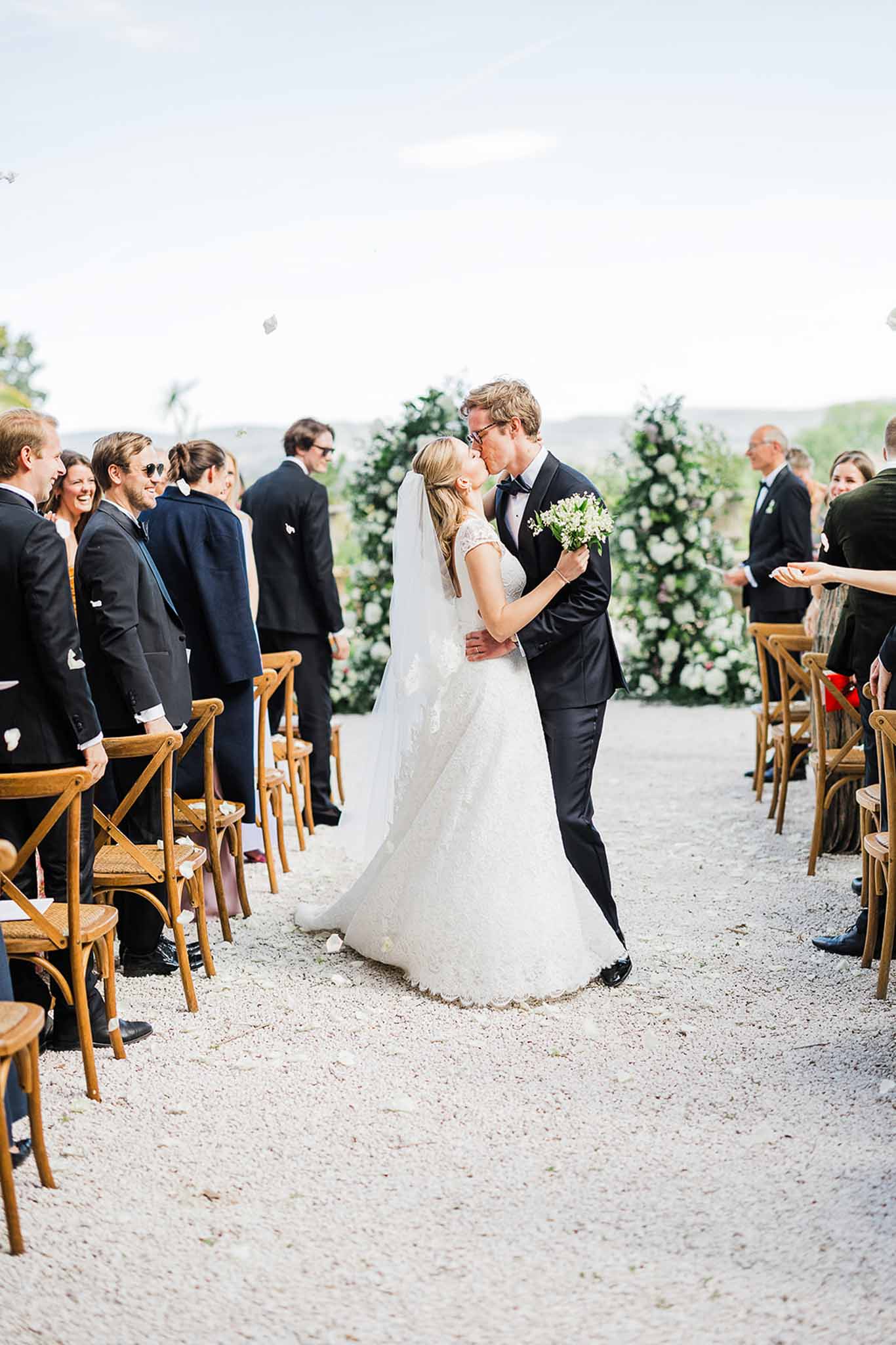 Bride and groom first kiss during outdoor wedding ceremony with guests and floral arrangements
