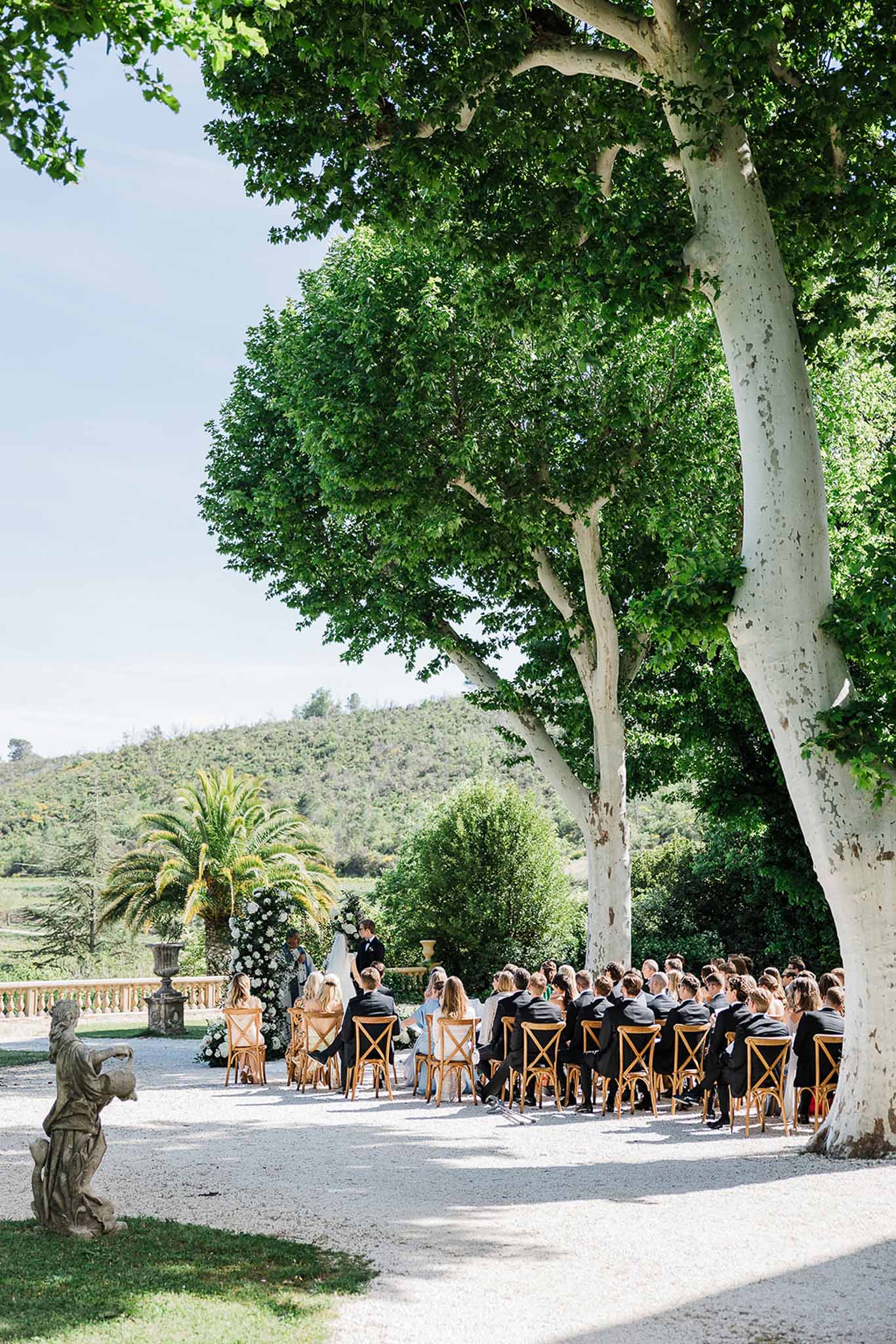 Wedding ceremony in outdoor courtyard with guests seated under large tree at Mediterranean estate
