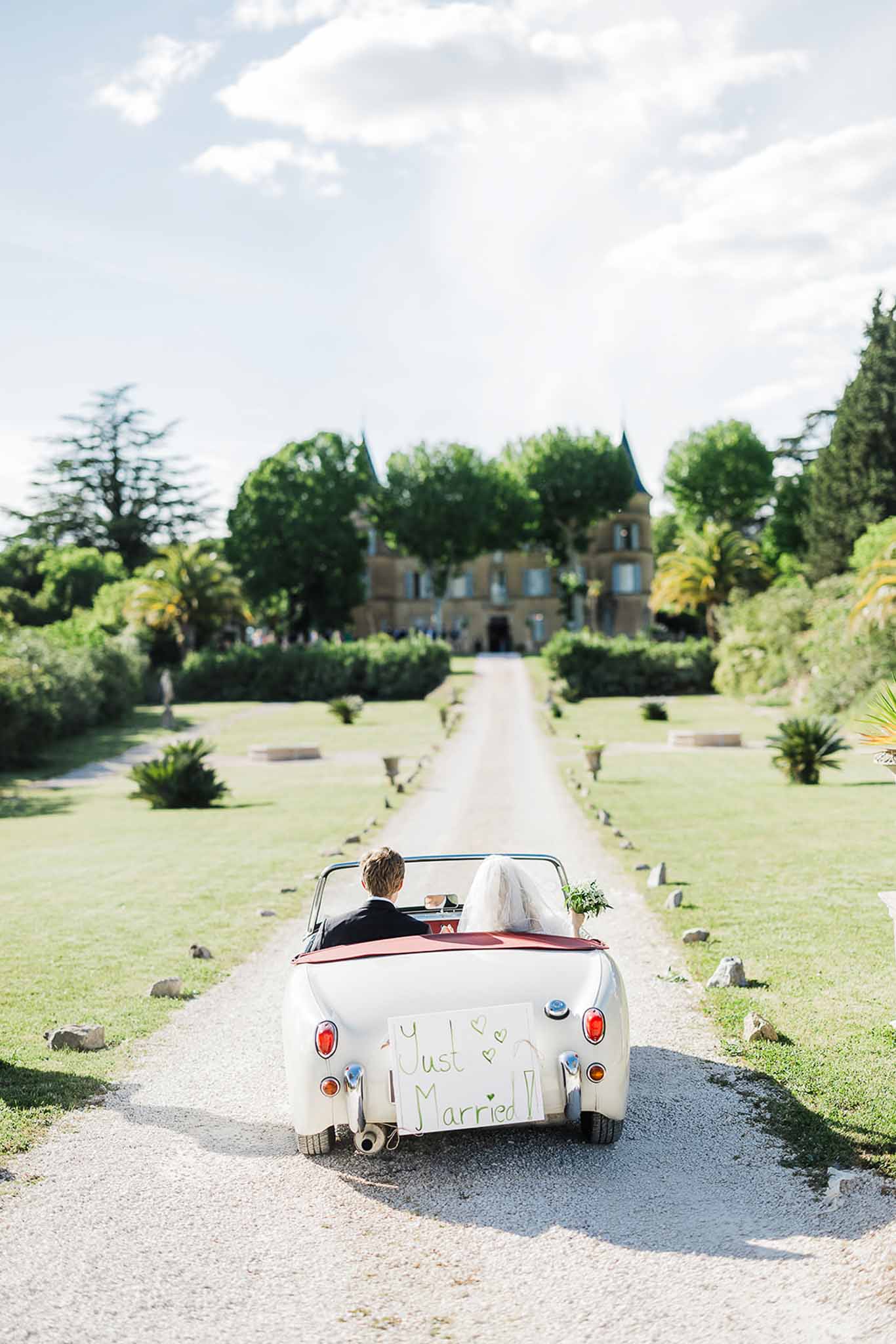Bride and groom driving vintage convertible down château driveway after wedding ceremony