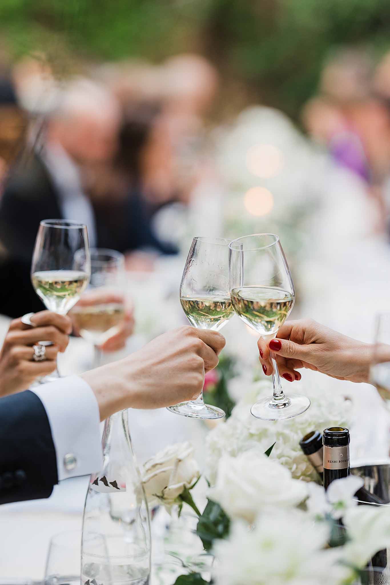Wedding guests raising wine glasses in toast at outdoor reception with white floral centerpieces
