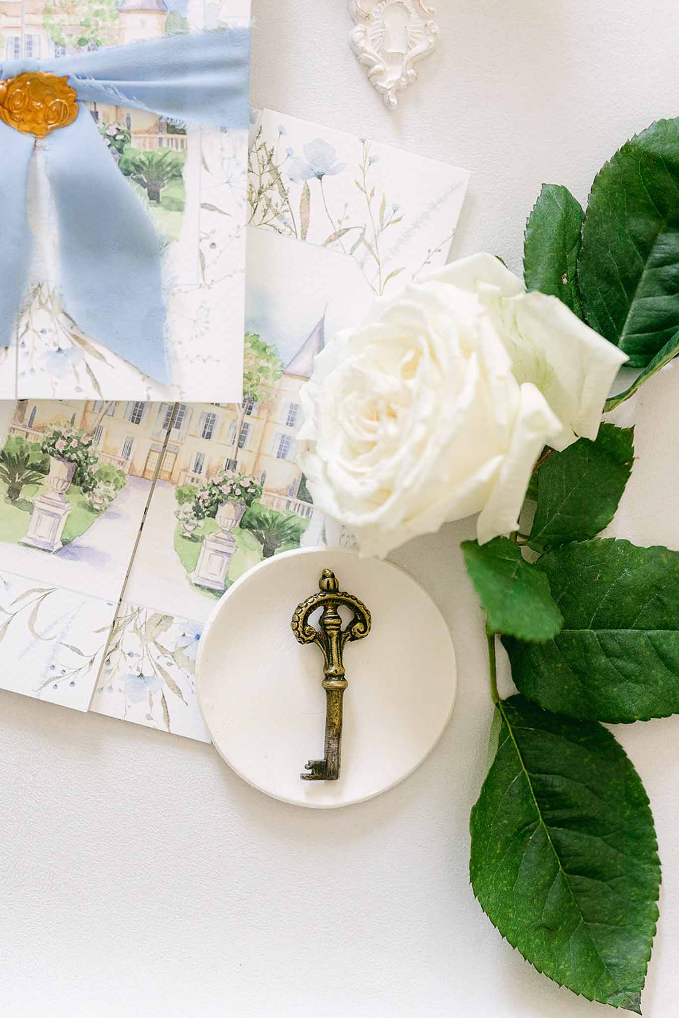 Wedding invitation flat lay with watercolor botanical designs, gold key, and cream rose on white surface