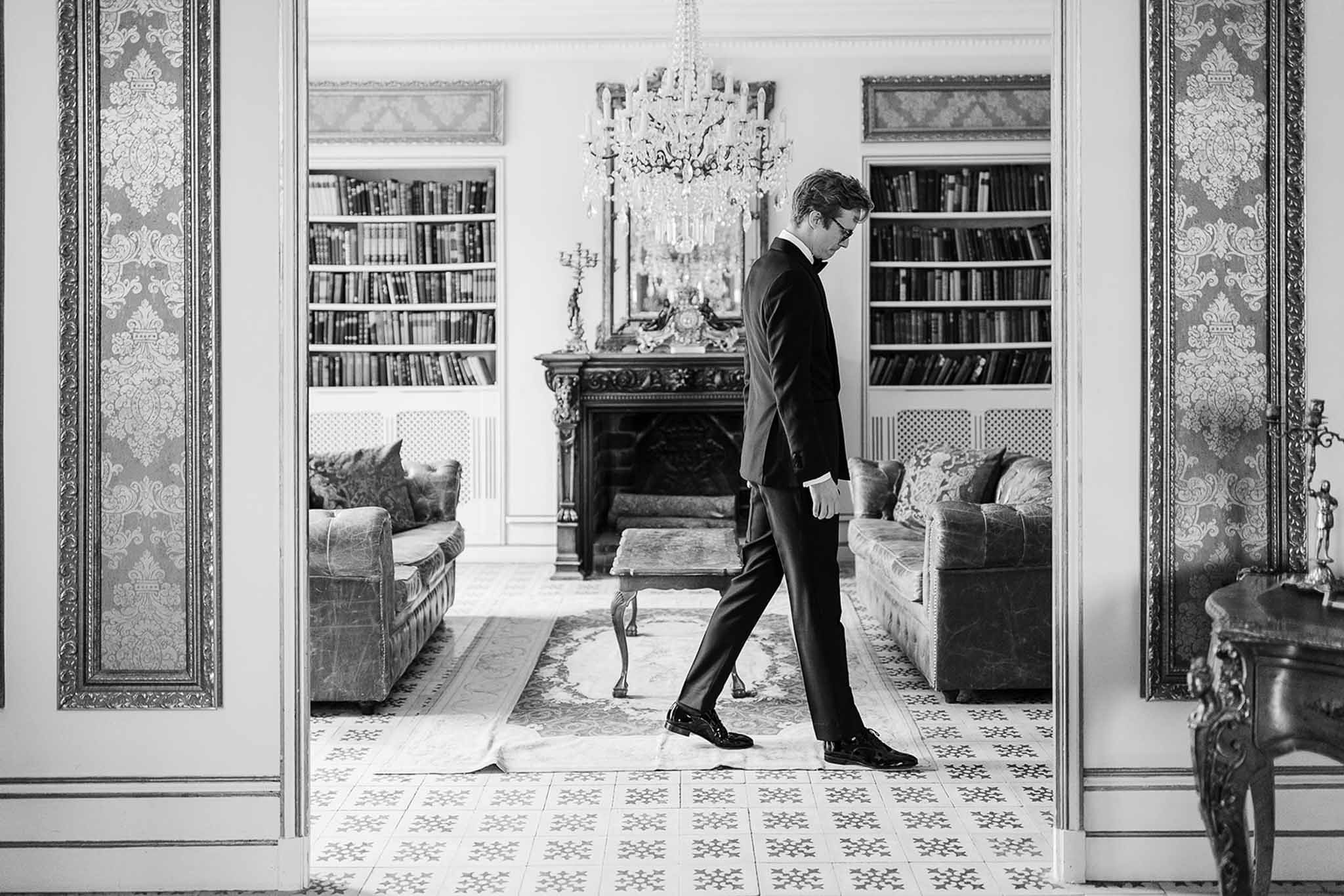 Groom in formal tailcoat walking through ornate library room with bookshelves and chandelier