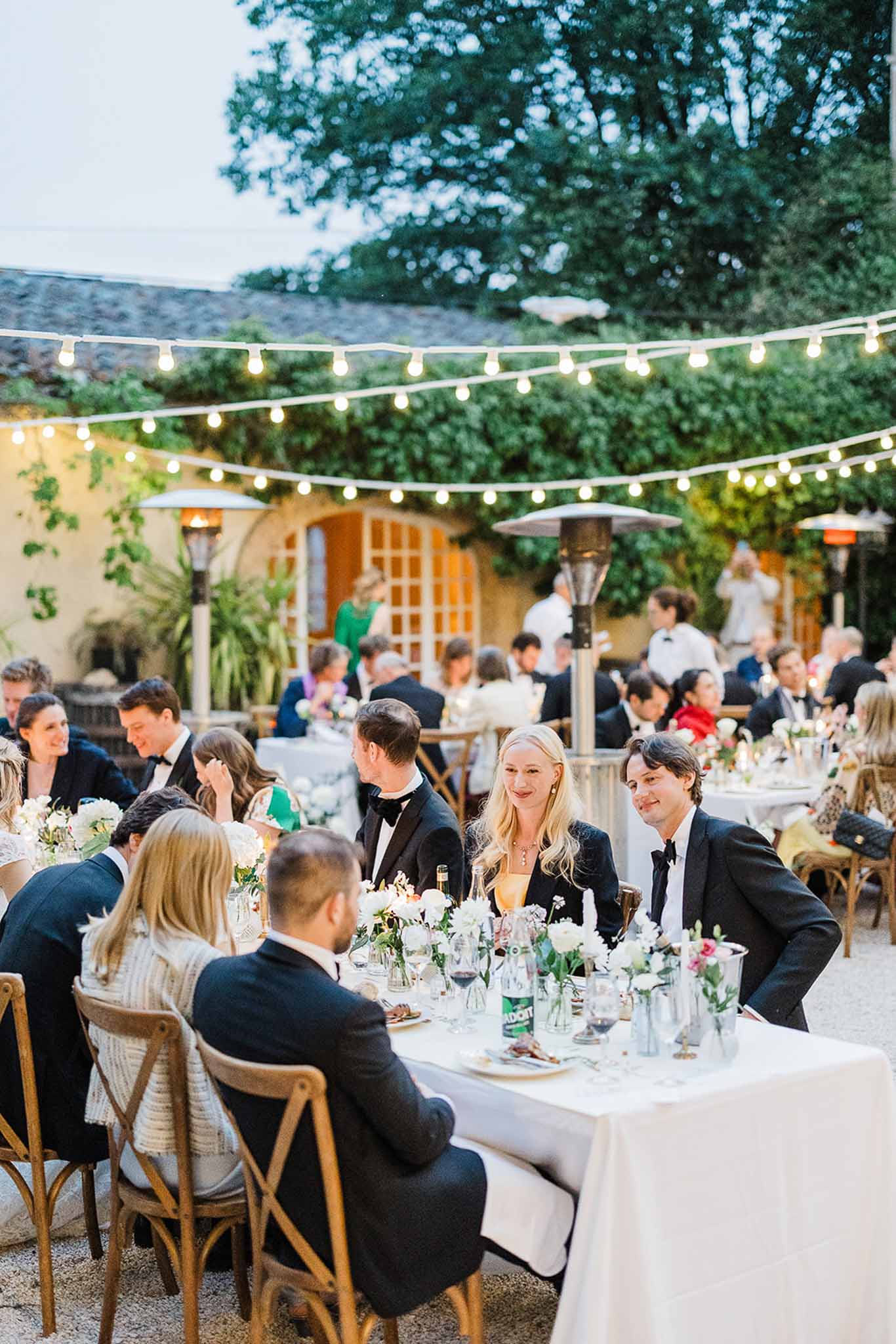 Outdoor reception dinner with guests seated at long tables in Mediterranean courtyard with string lighting