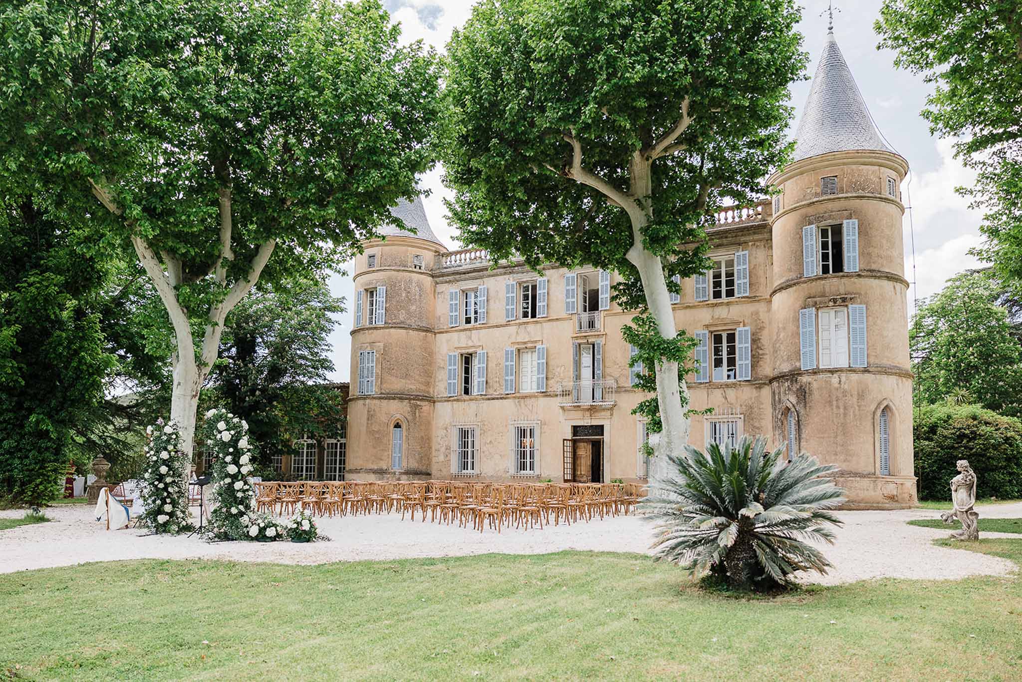 Outdoor ceremony setup in stone château courtyard with wooden chairs and floral arch