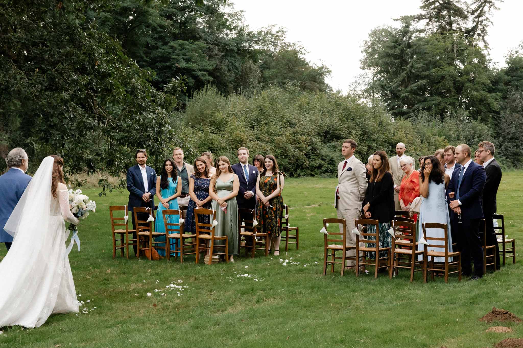 Outdoor wedding ceremony with couple and guests in tree-lined garden setting with wooden chairs