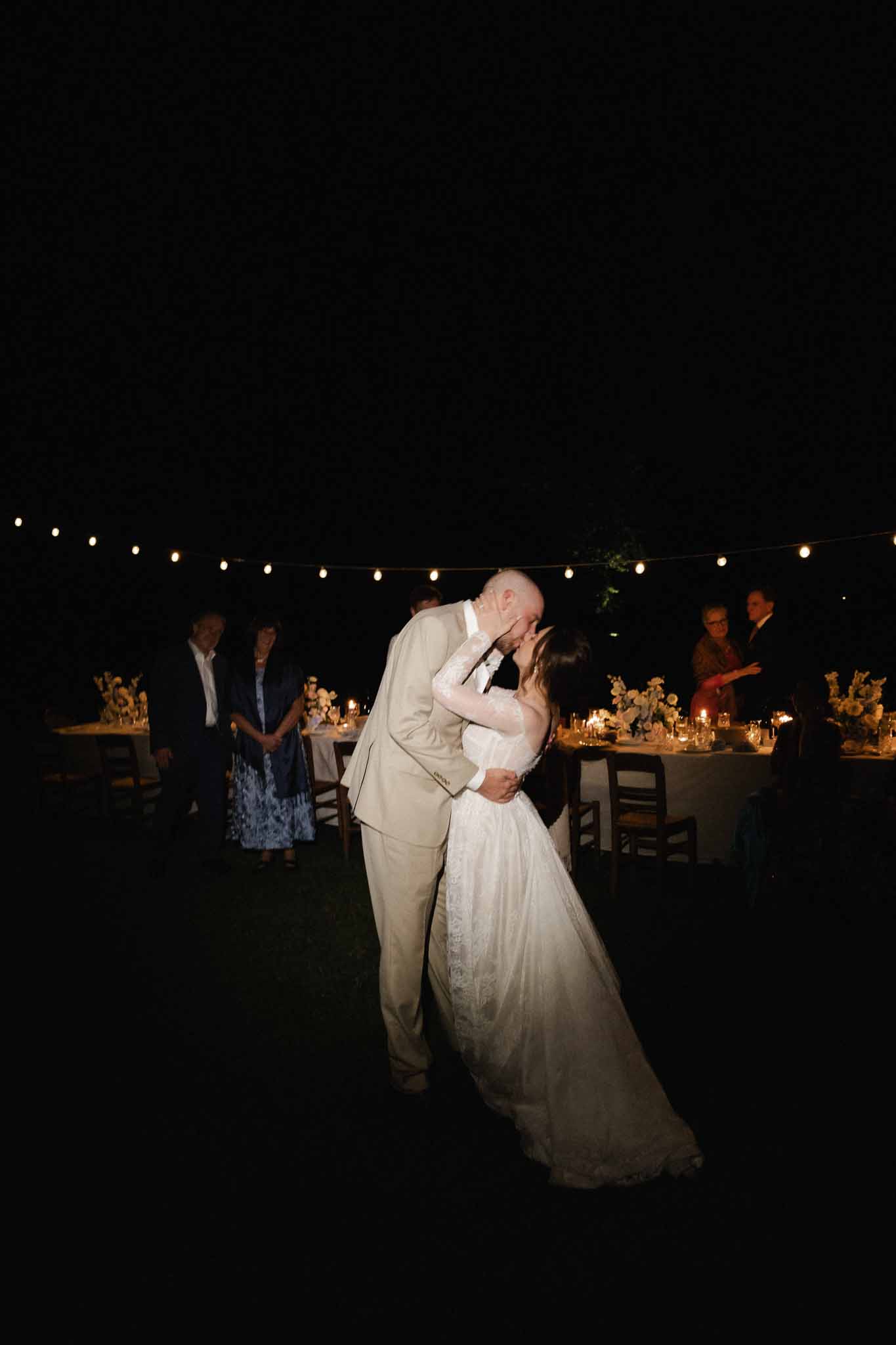 Bride and groom first dance at outdoor nighttime wedding reception with string lights and guests watching