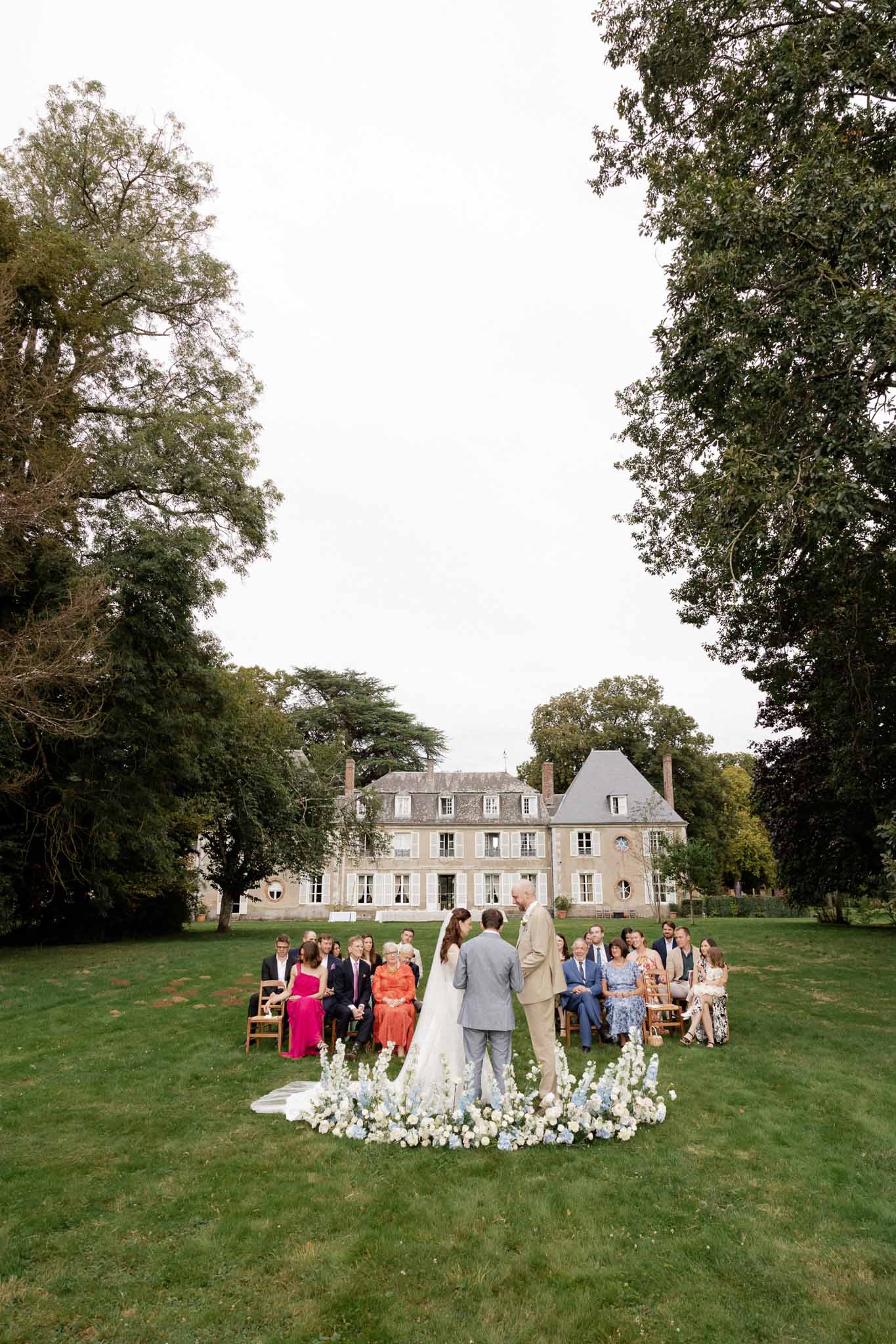 Wedding ceremony with circular floral arch on château lawn with guests in wooden chairs