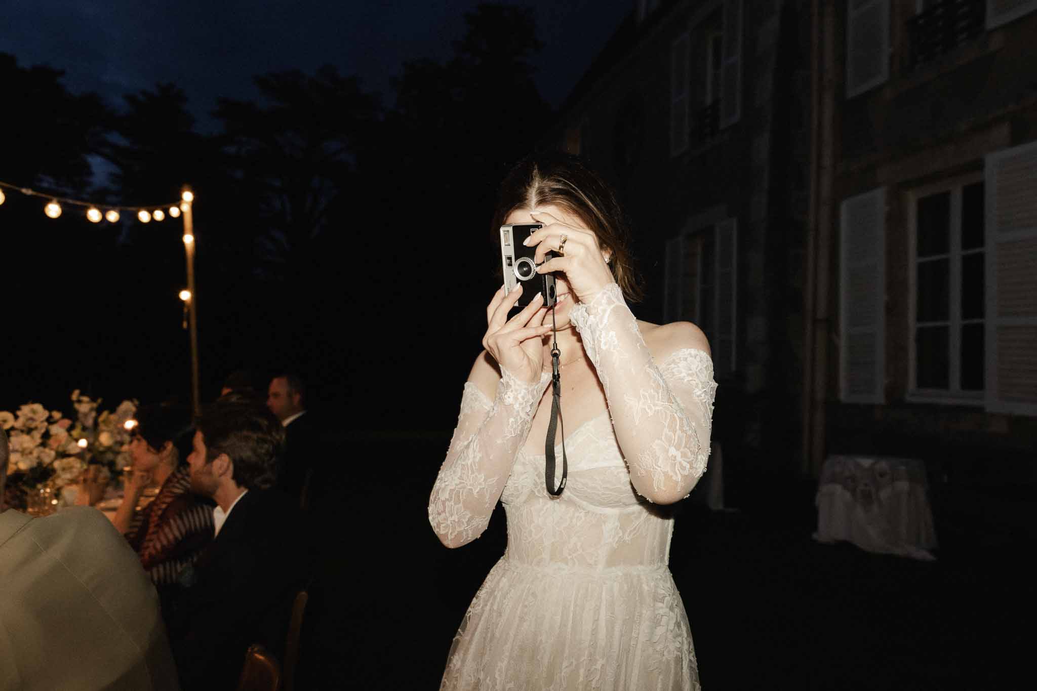 Bride with vintage camera during evening reception in outdoor courtyard with string lights and guests