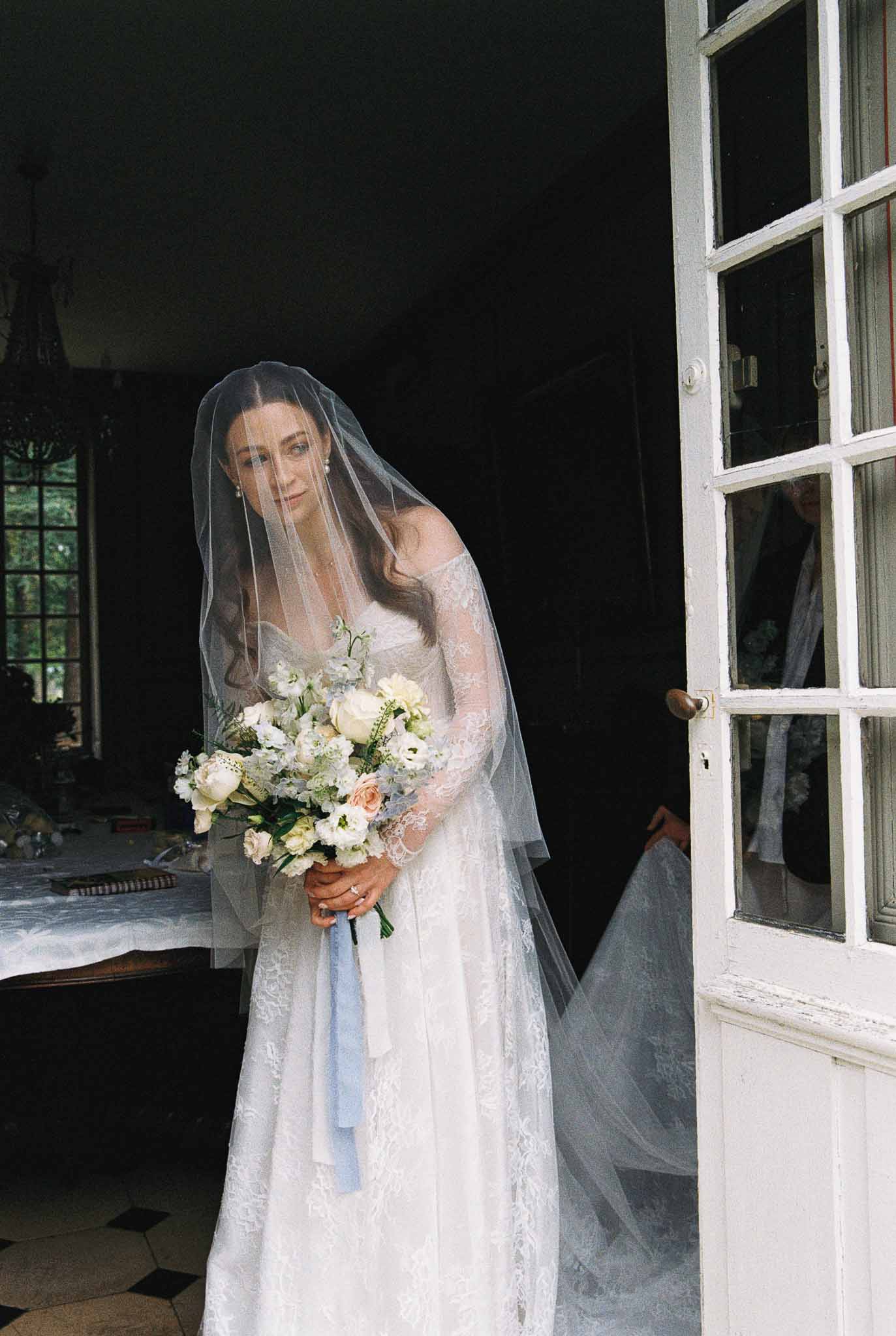 Bride in ivory lace dress holding bouquet near French door at indoor wedding venue