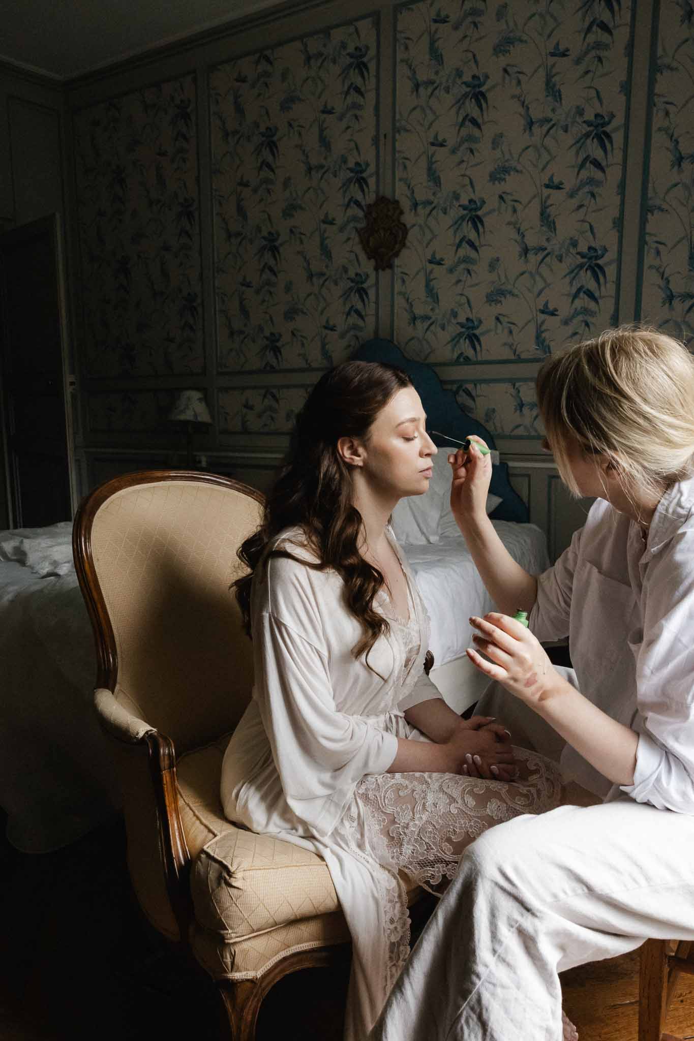Bride receiving makeup application while sitting in elegant chair during wedding preparation