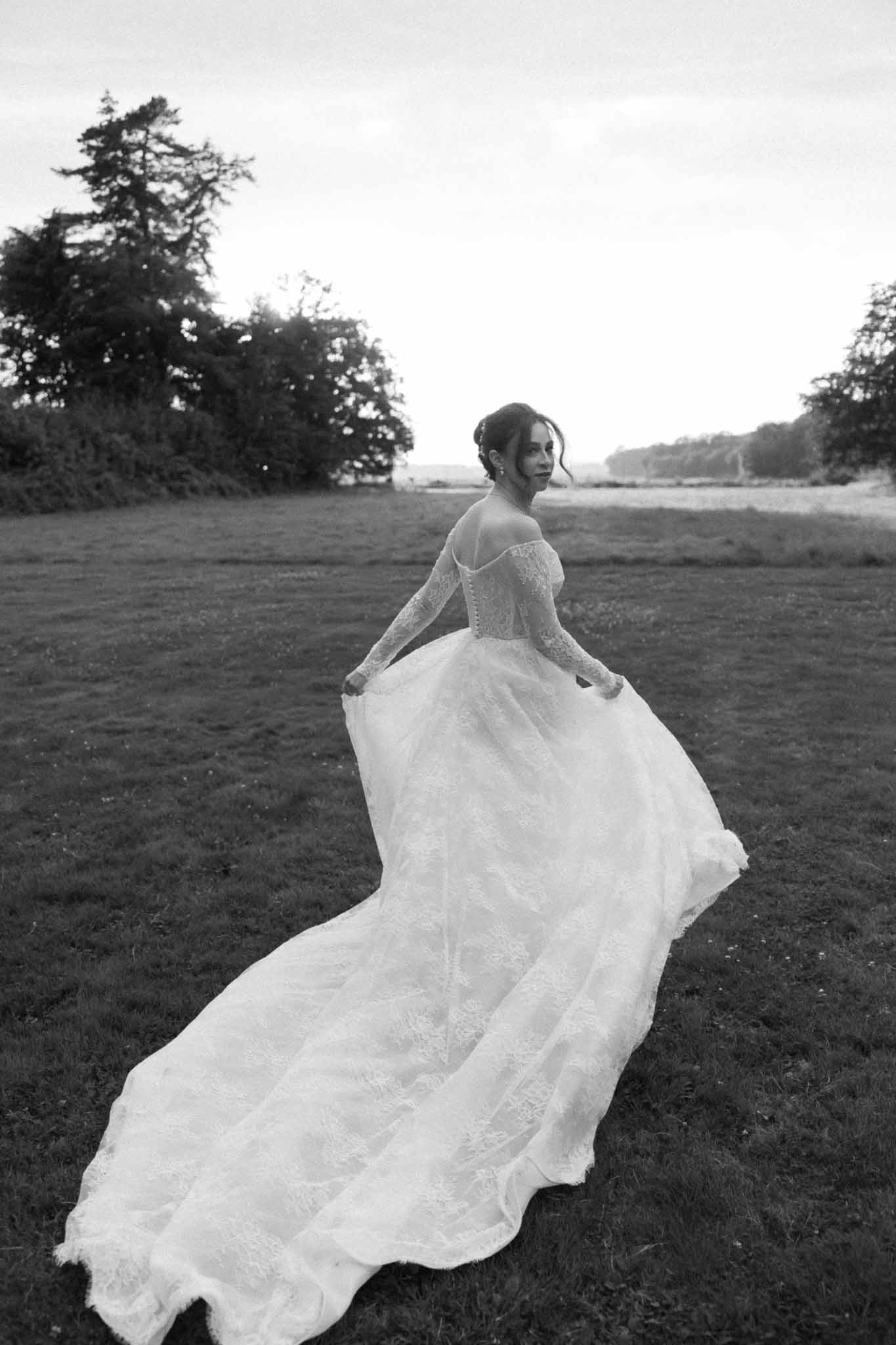Bride in flowing lace wedding dress walking through open field with water in distance