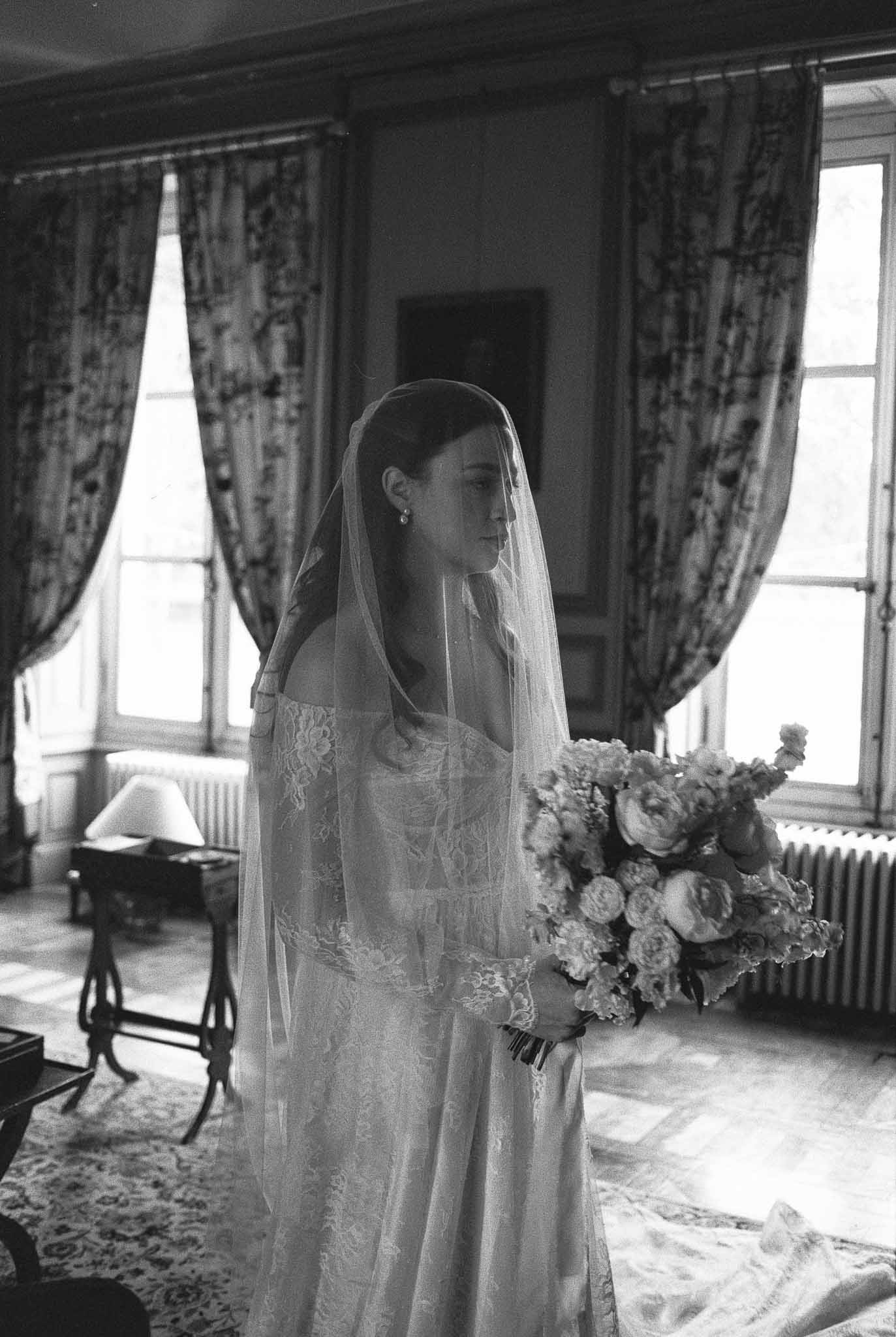 Bride in lace wedding dress with veil holding bouquet in elegant interior room with tall windows