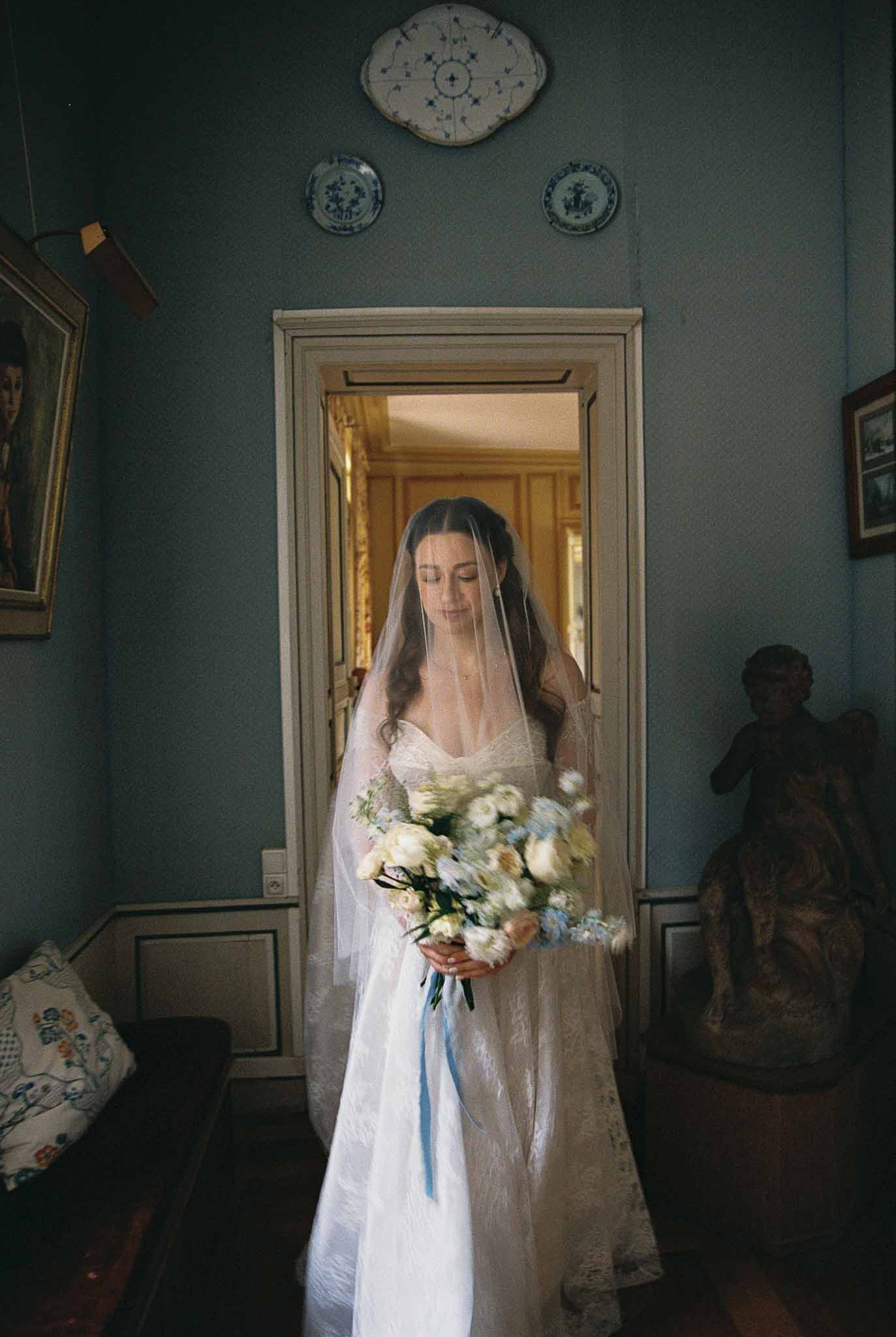 Bride in white slip dress holding blue and white bouquet standing in doorway with teal walls and ceramic plates