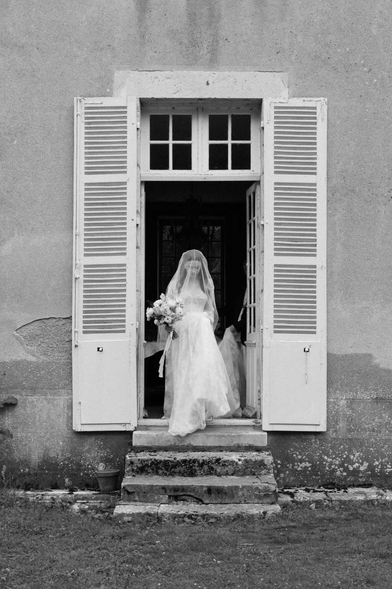 Bride in wedding dress and veil standing in stone building doorway with white shutters