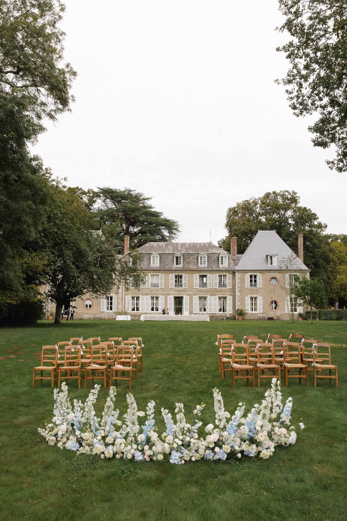 Outdoor wedding ceremony setup on château lawn with floral installation and neoclassical French mansion backdrop