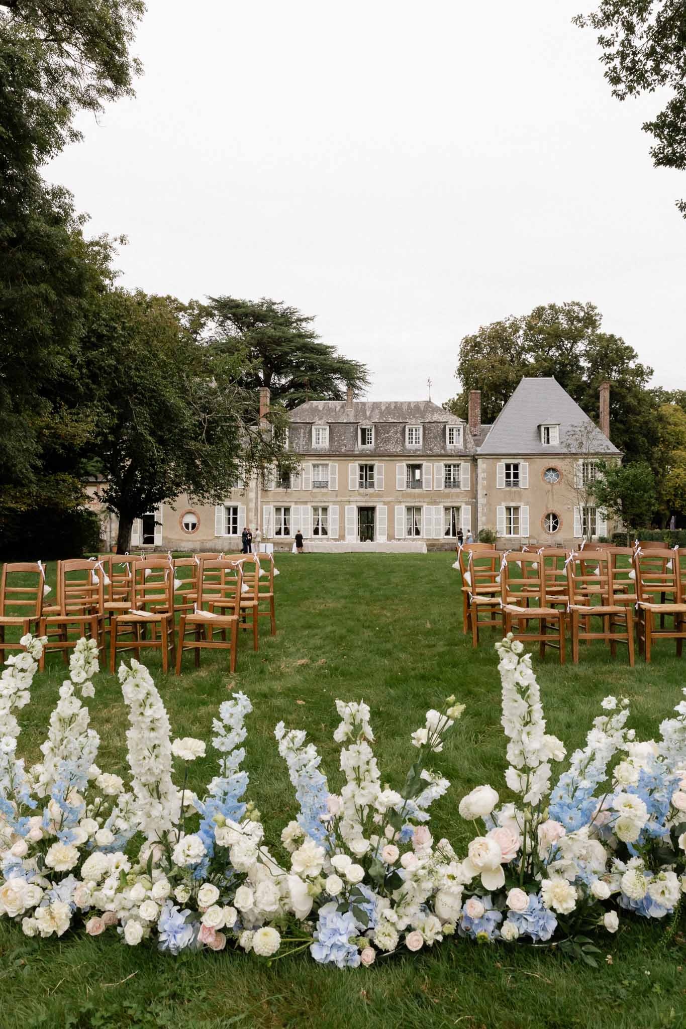 Outdoor wedding ceremony setup on lawn of French château with floral aisle borders and cross-back chairs