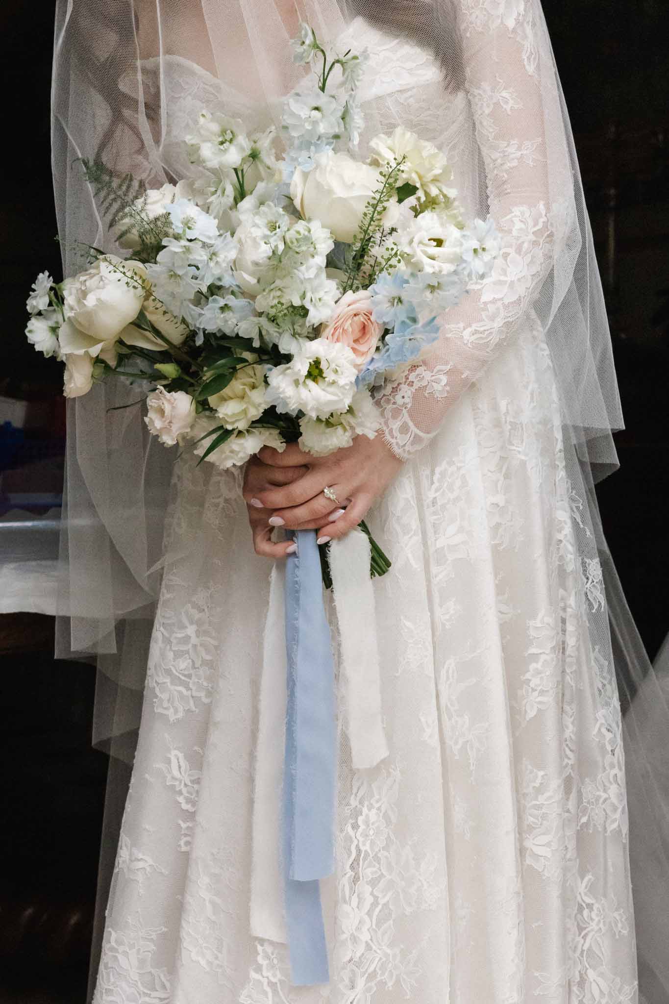 Bride holding white and blue wedding bouquet in ivory lace dress with long sleeves and embroidered veil