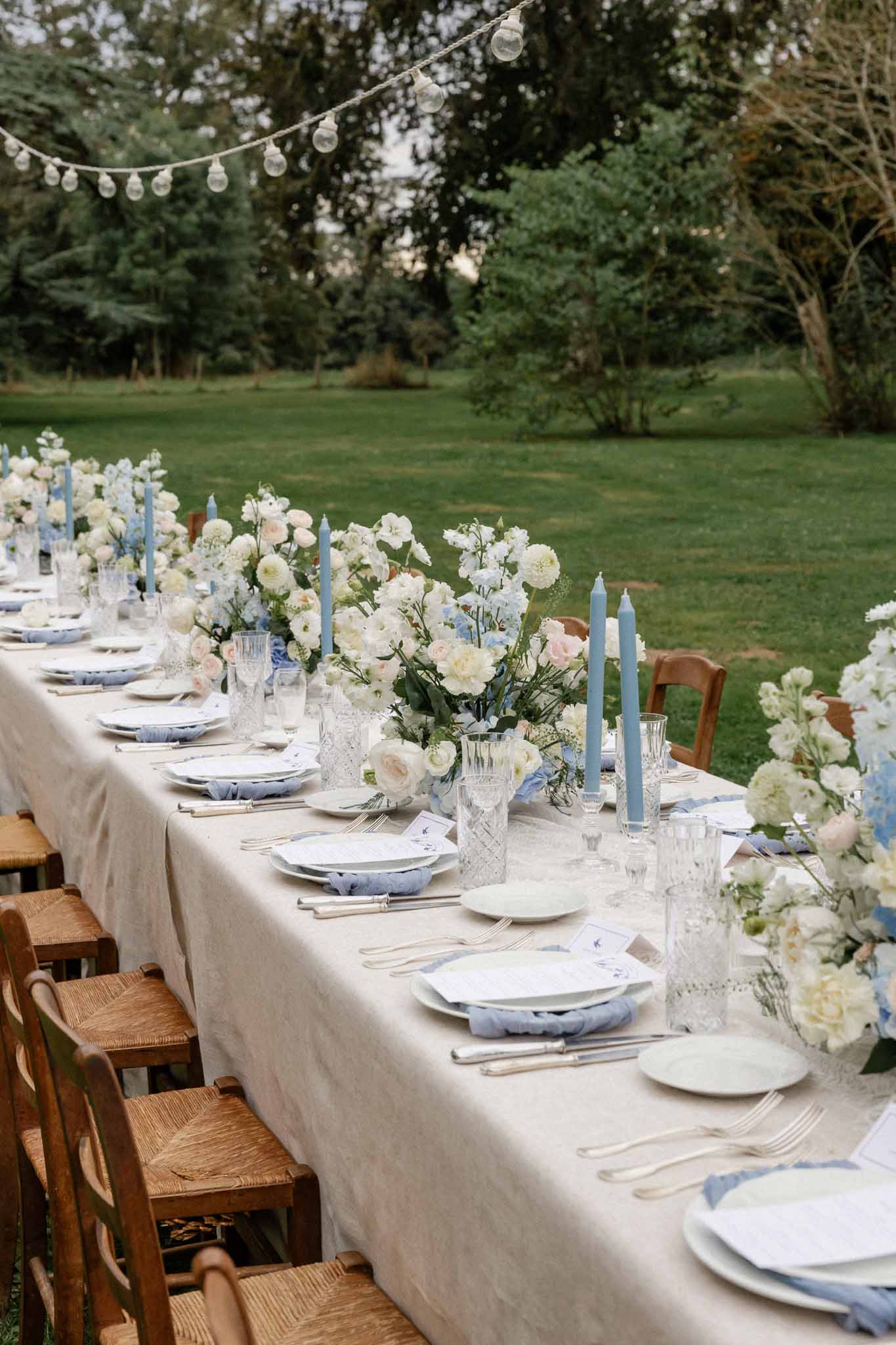 Outdoor reception tablescape with ivory linens and blue napkins on manicured lawn with string lighting