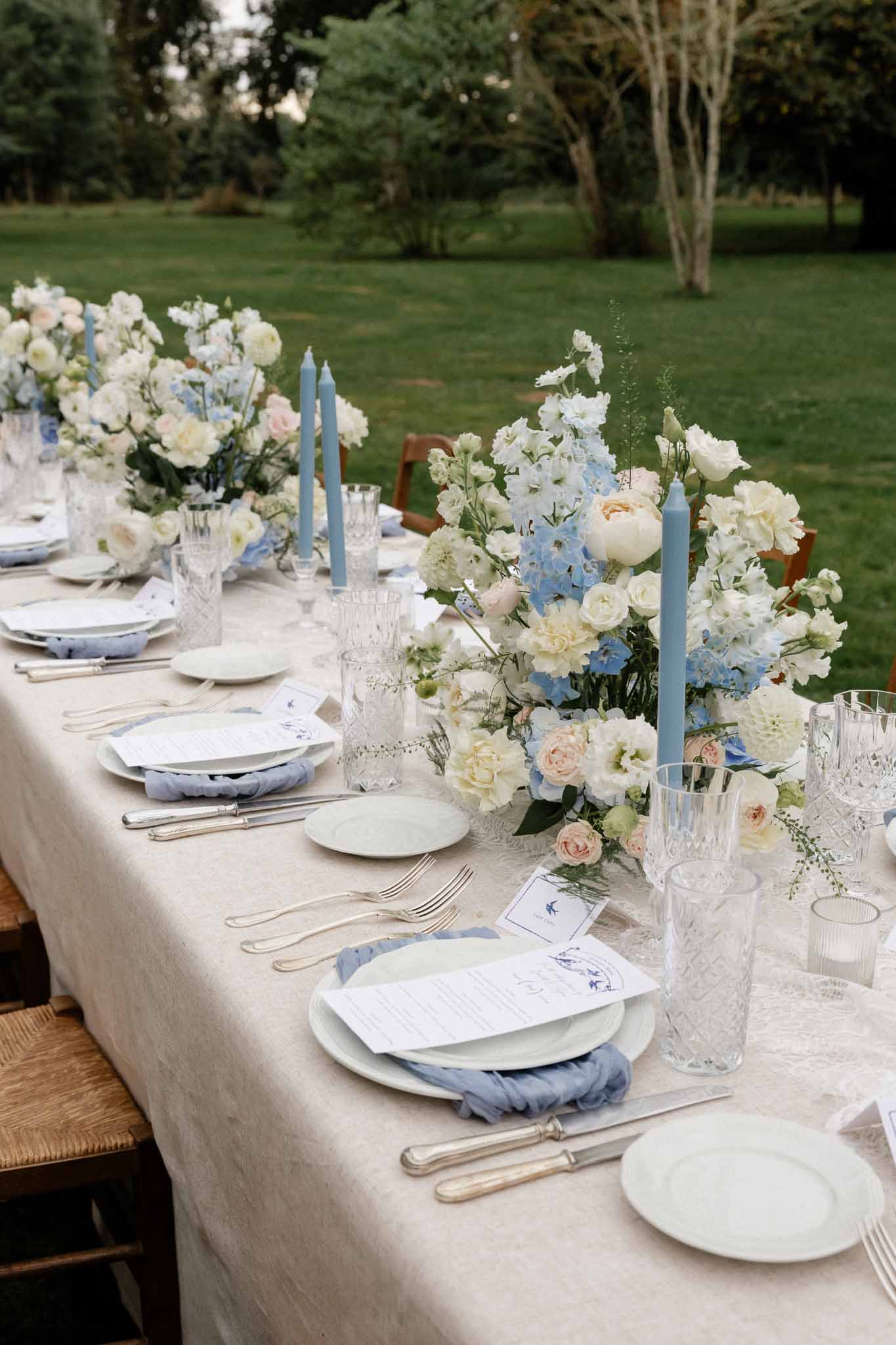 Outdoor wedding reception table setting with ivory linens and blue floral centerpieces in garden venue