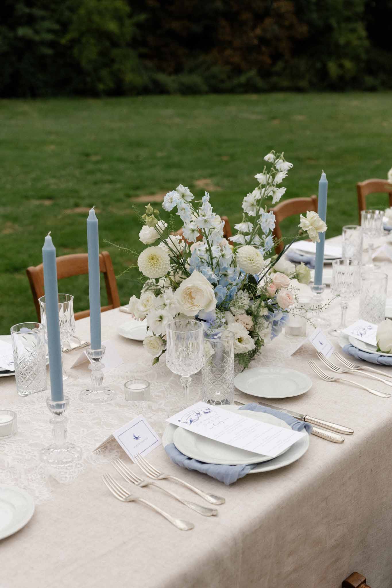 Elegant outdoor wedding reception tablescape with ivory linens and blue accents on manicured lawn