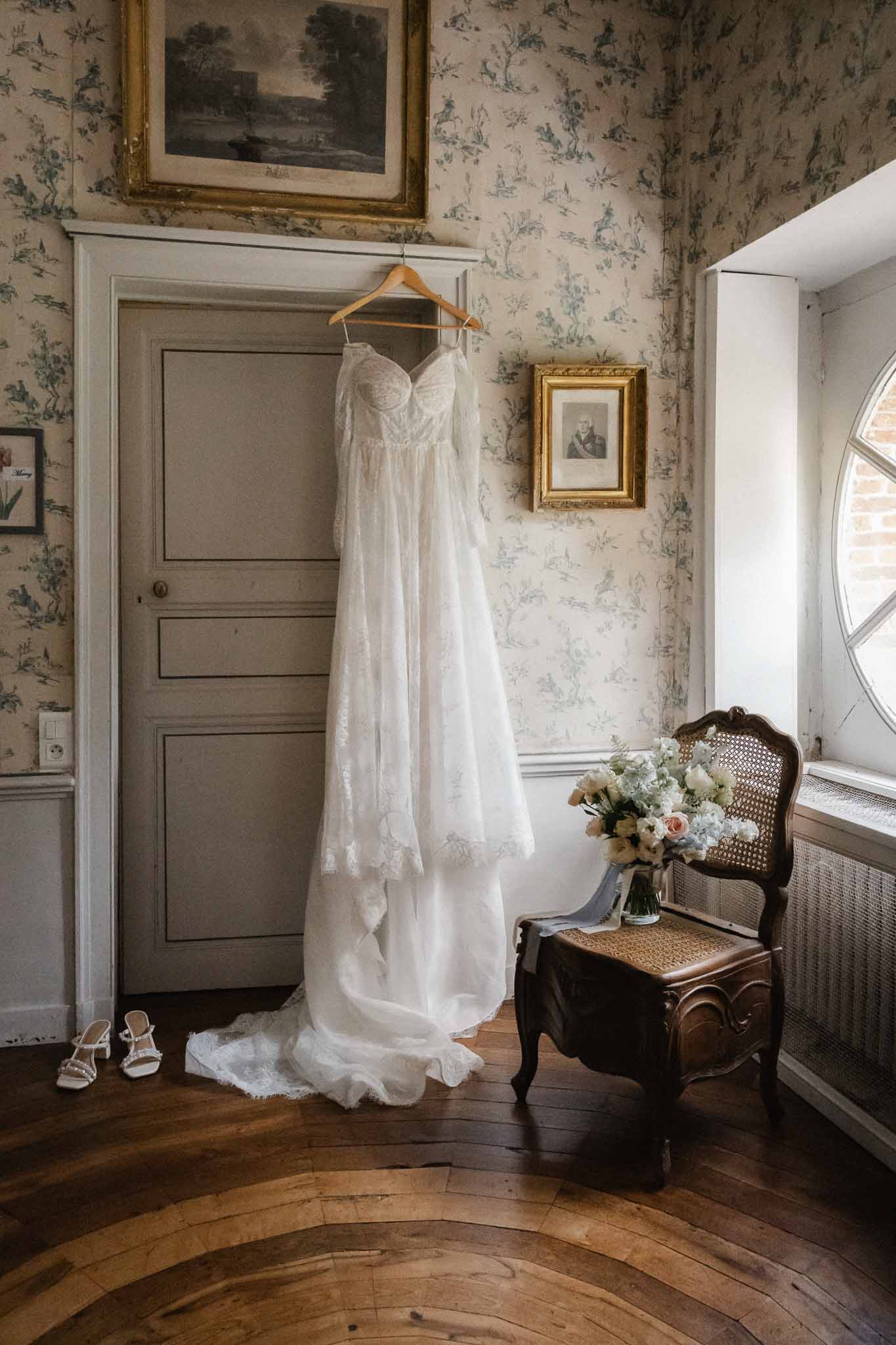 Wedding dress and bridal bouquet hanging in classical bedroom with toile wallpaper