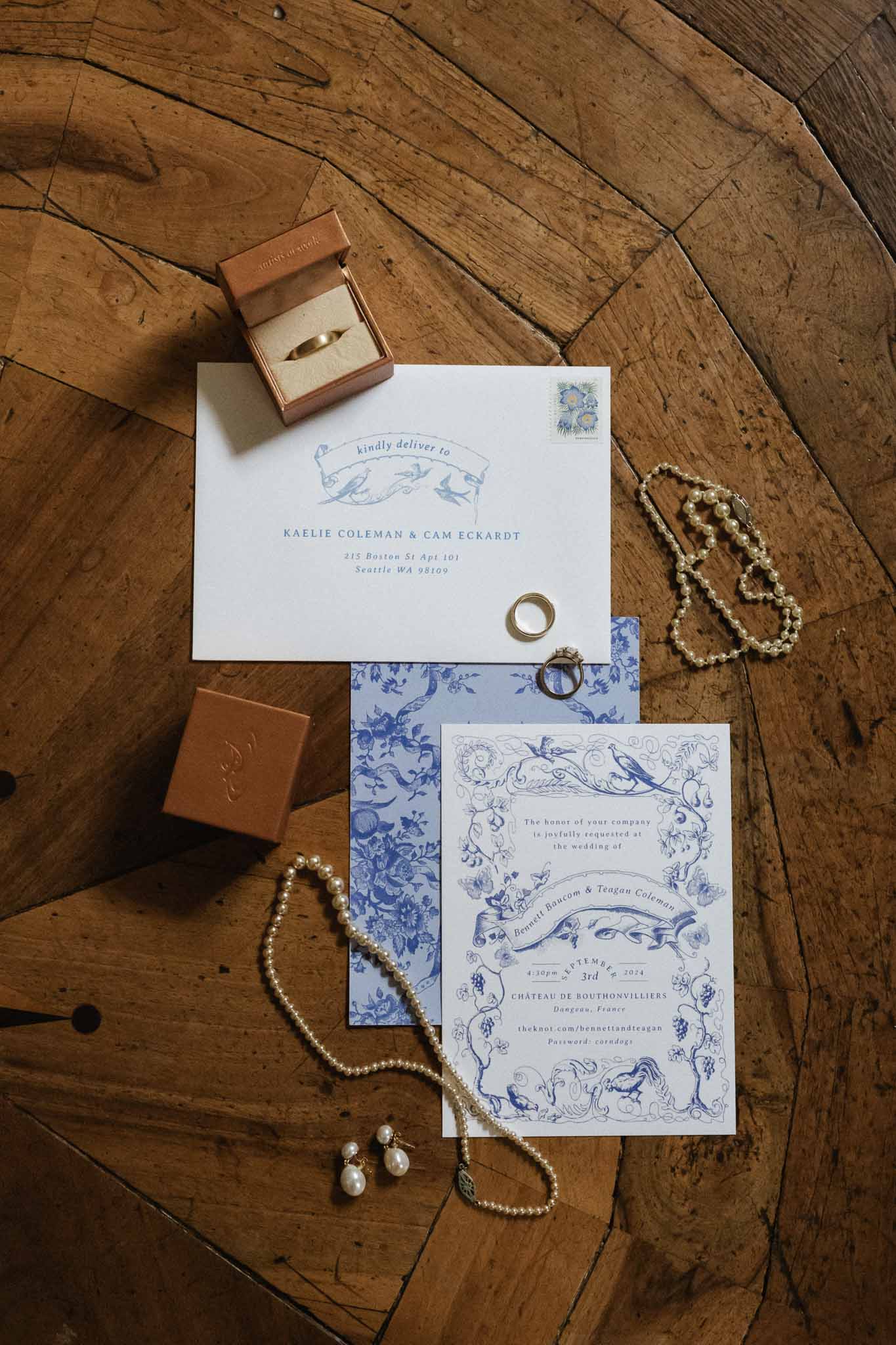 Wedding invitation flat lay with blue botanical stationery, gold rings, and pearl jewelry on wooden floor