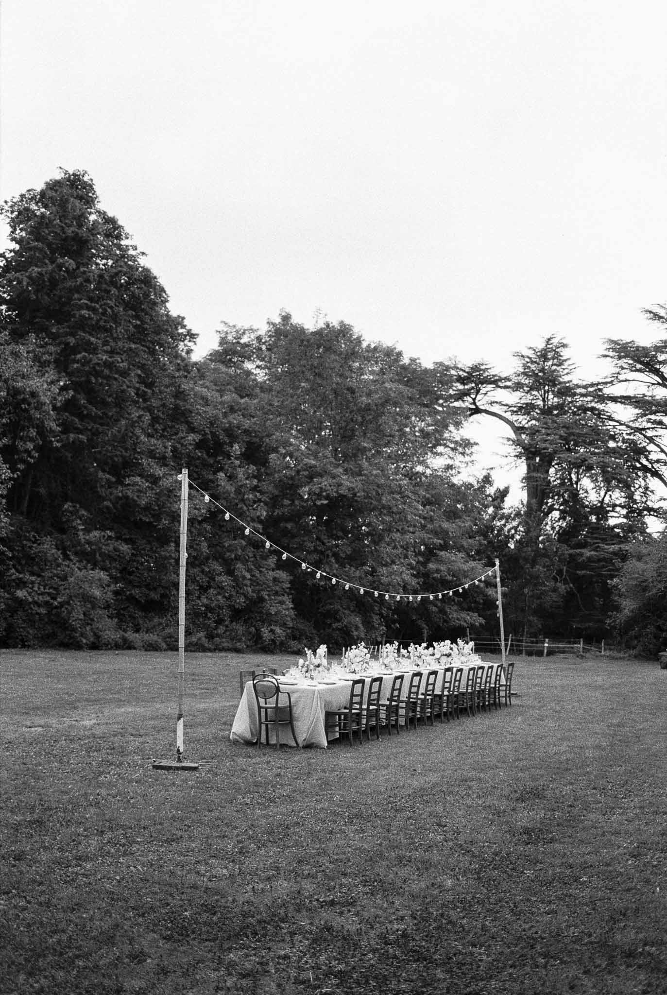 Outdoor reception table setup with string lights in open field surrounded by trees