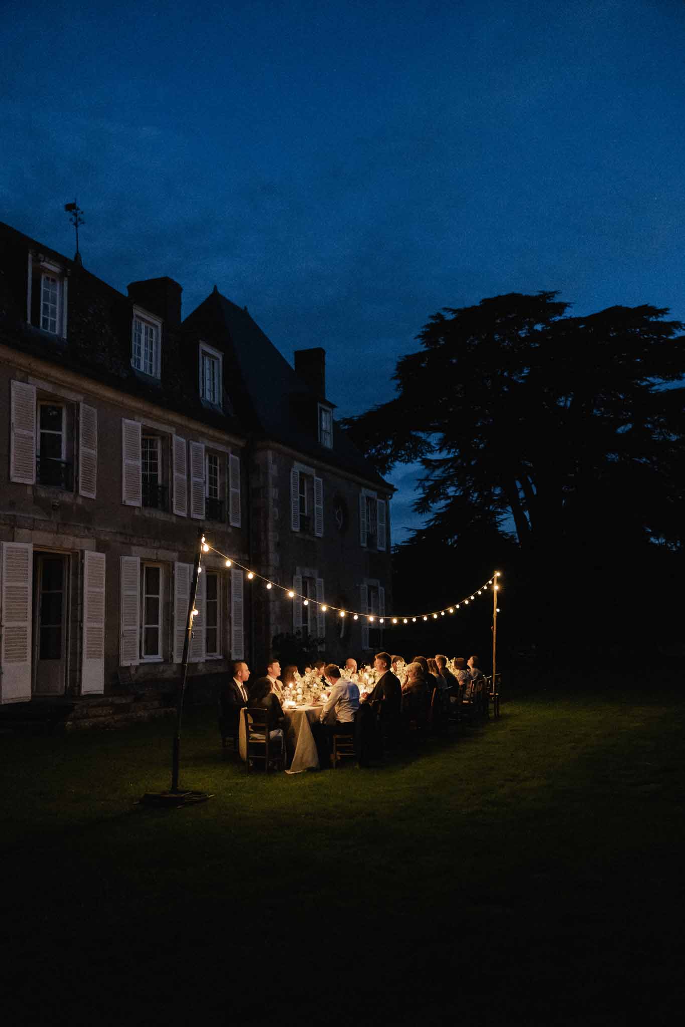 Evening reception dinner on manor house grounds with guests seated at long table under string lights