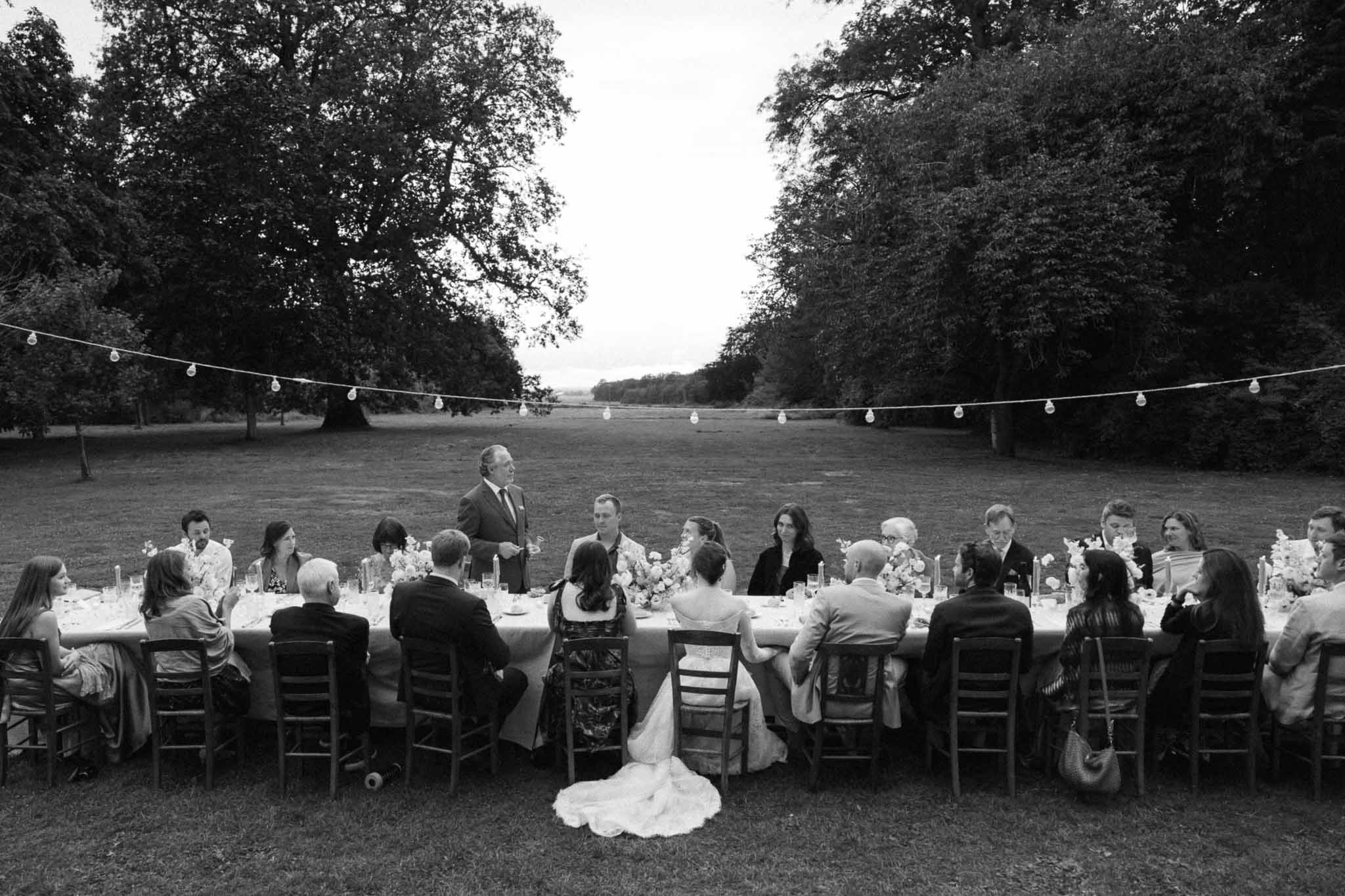 Outdoor reception dinner with guests seated at long table under string lights in parkland setting