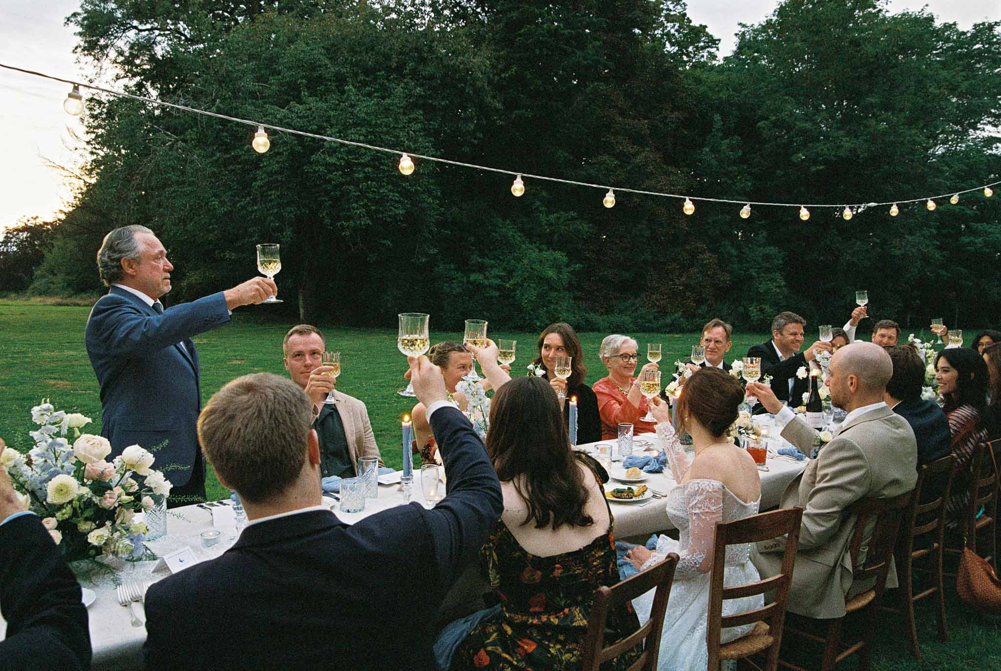 Wedding reception speeches with guests raising glasses at outdoor dinner table with string lights