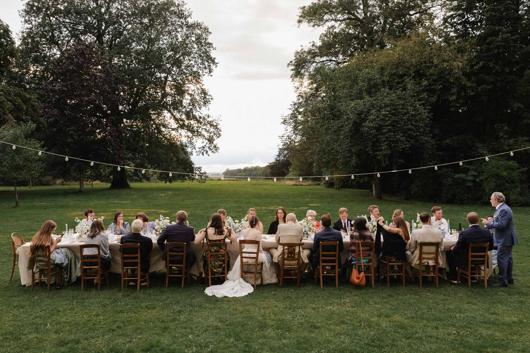 Outdoor wedding reception with guests at long table in parkland setting with string lights