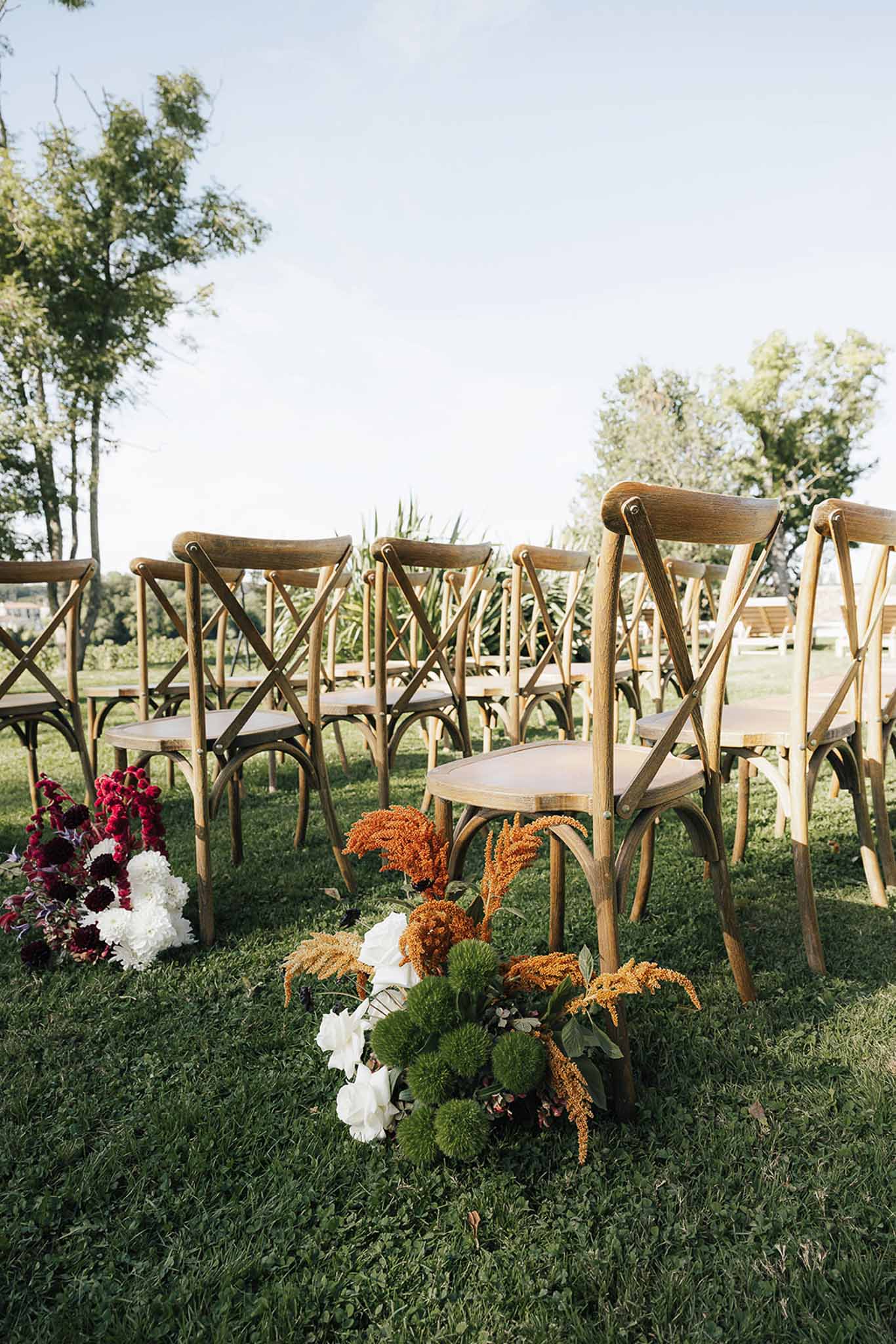 Ceremony aisle with burnt orange amaranthus and burgundy dahlia ground arrangements beside cross-back chairs