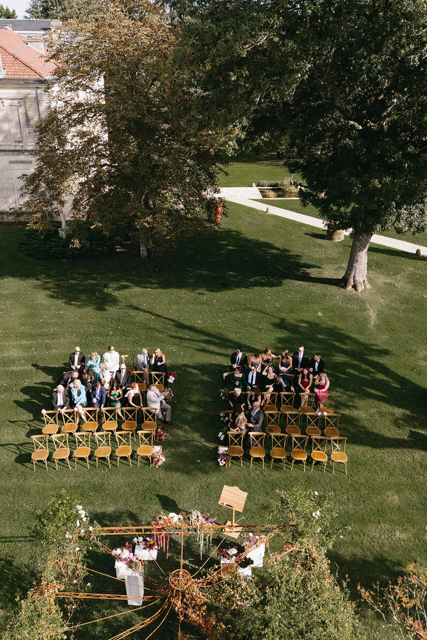 Aerial view of outdoor garden wedding ceremony setup with geometric floral arch and cross-back chairs on manicured lawn