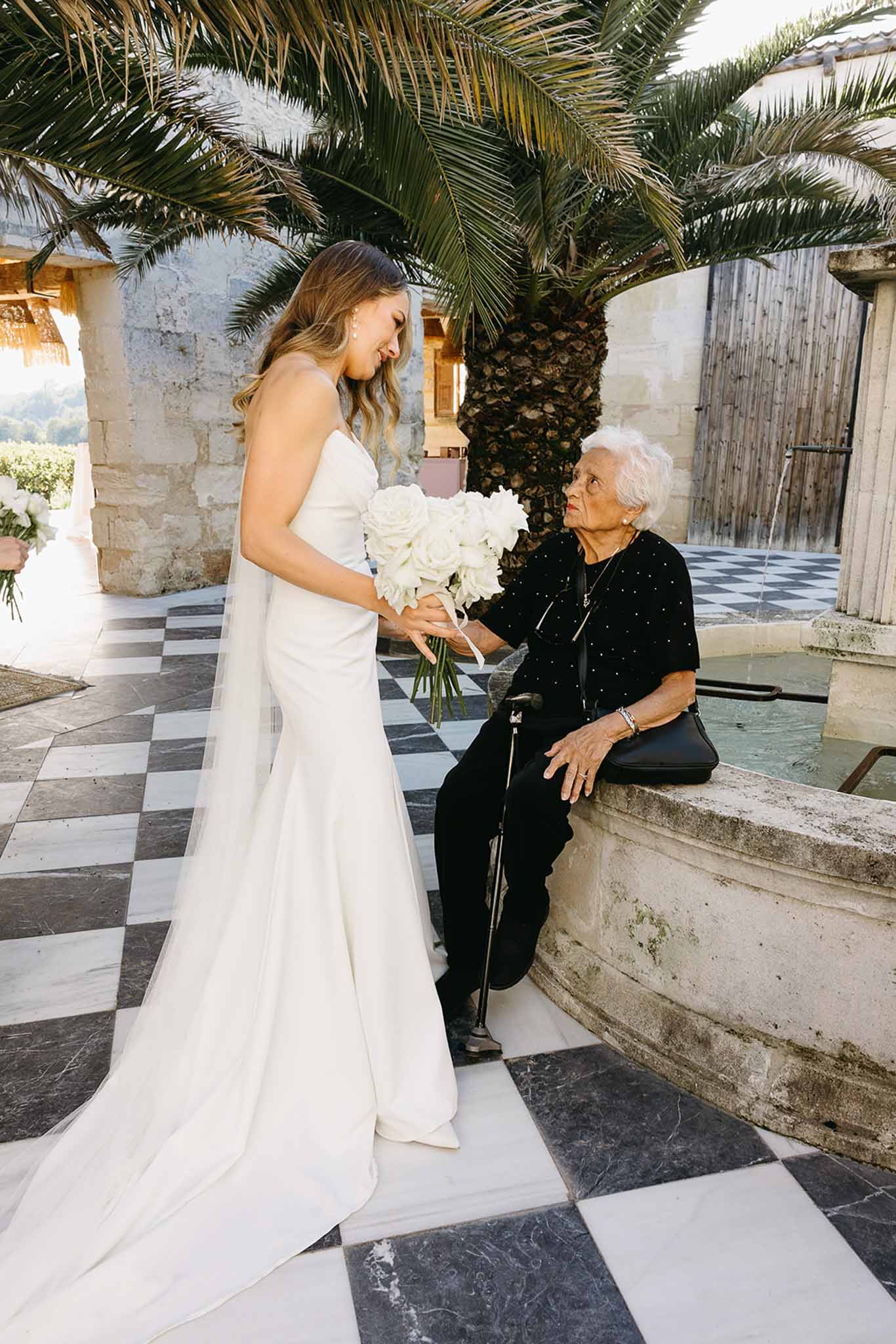 Bride in ivory dress with elderly woman in Mediterranean courtyard with palm trees and stone architecture