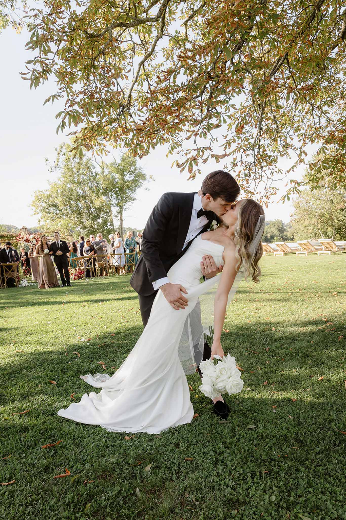 Groom dips and kisses bride during outdoor garden reception with guests watching from gold chairs