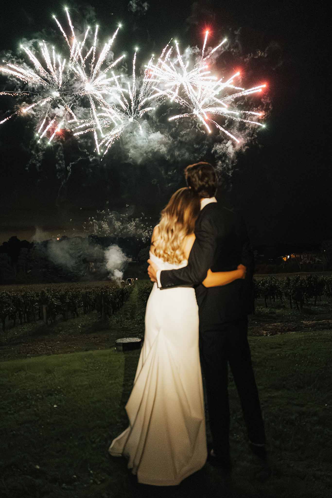 Bride and groom watching fireworks display on outdoor lawn during wedding reception exit celebration