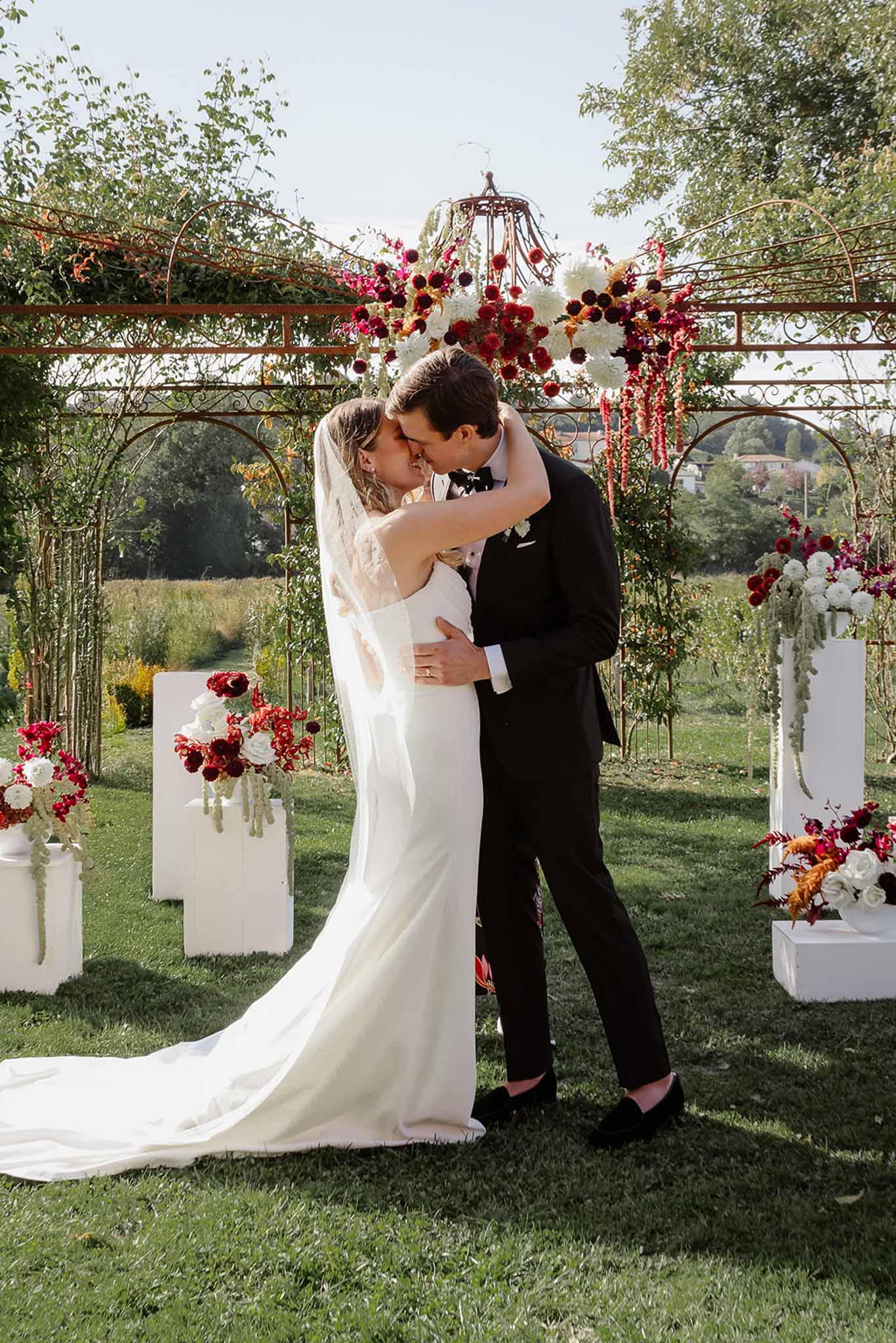 Bride and groom first kiss during outdoor garden ceremony with floral arch and dramatic hanging installation