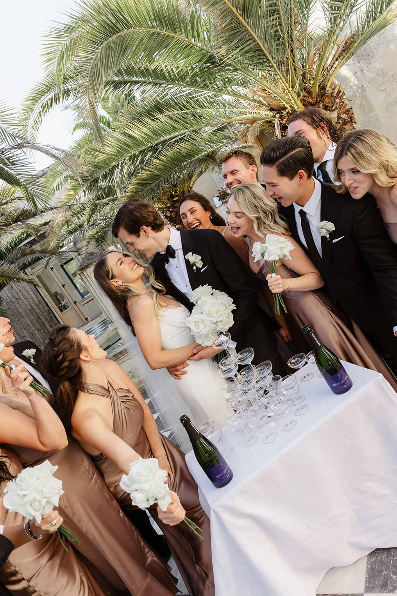 Bride and groom with bridal party gathered around cocktail table in outdoor courtyard with palm trees